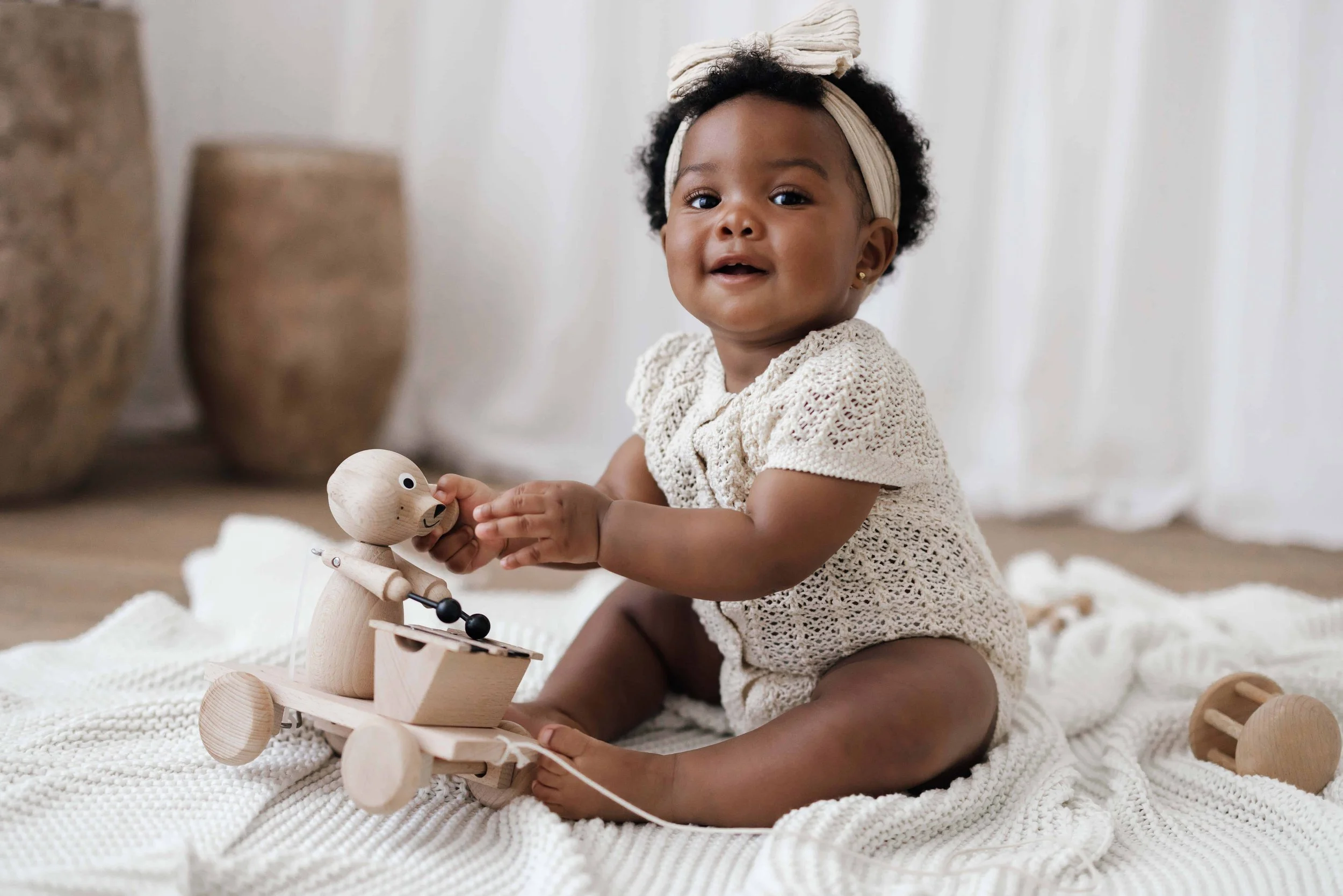 A baby girl with curly hair, wearing a beige knit dress and a head wrap, sitting on a white rug and playing with a wooden toy motorcycle.