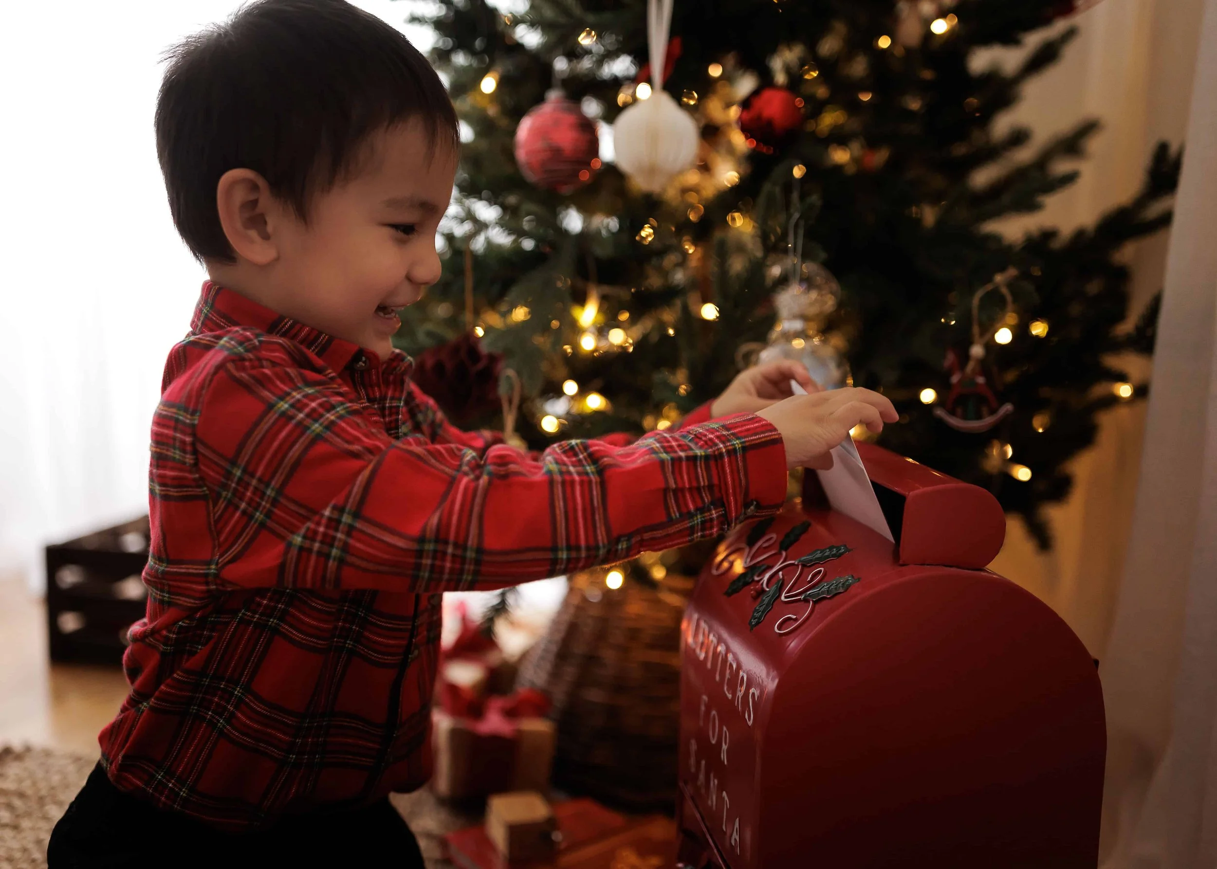 A young boy in a red plaid shirt placing a letter in a red Christmas mailbox in front of a decorated Christmas tree with lights and ornaments.