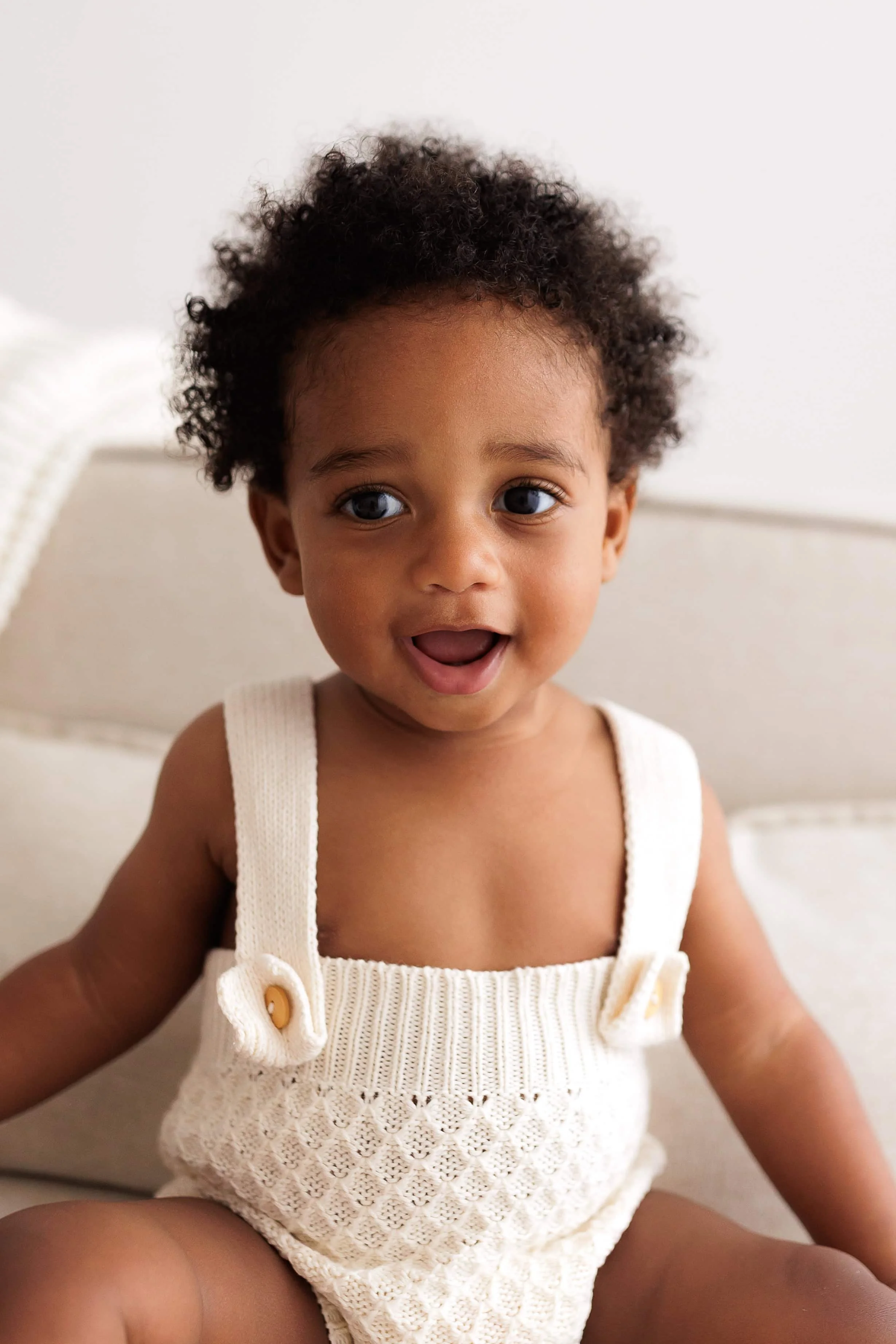 A happy young child with curly hair smiling and sitting on a light-colored couch.