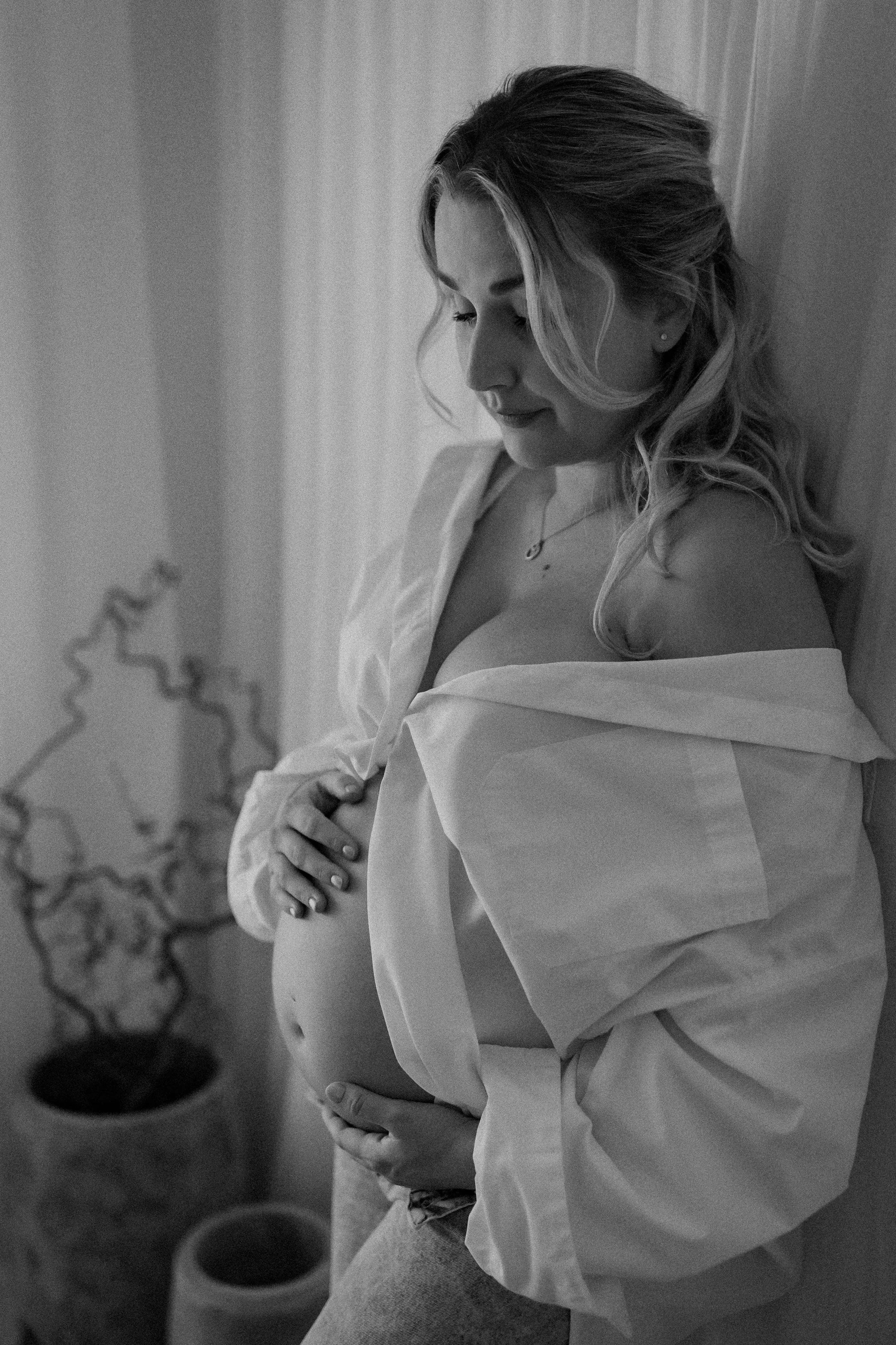 A pregnant woman with long wavy hair sitting down, wearing a loose shirt that falls off one shoulder. She is gently holding her pregnant belly with both hands, looking down at it softly. The background includes a decorative plant with tendrils.