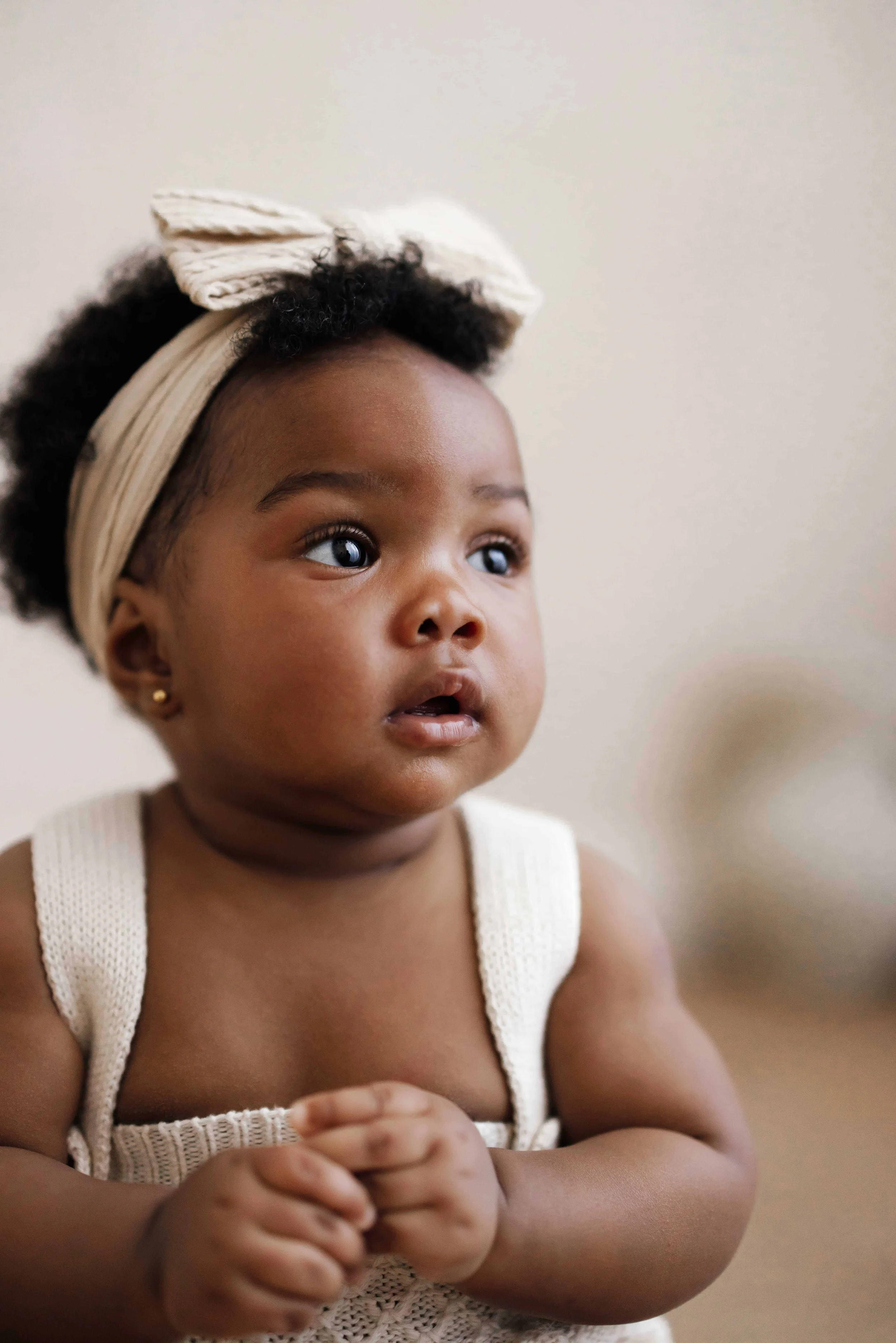 Close-up of a 8 month old girl with curly hair, wearing a beige headband with a bow, a cream-colored sleeveless top, and small gold earrings. She has wide eyes and an attentive expression, with her hands clasped together.