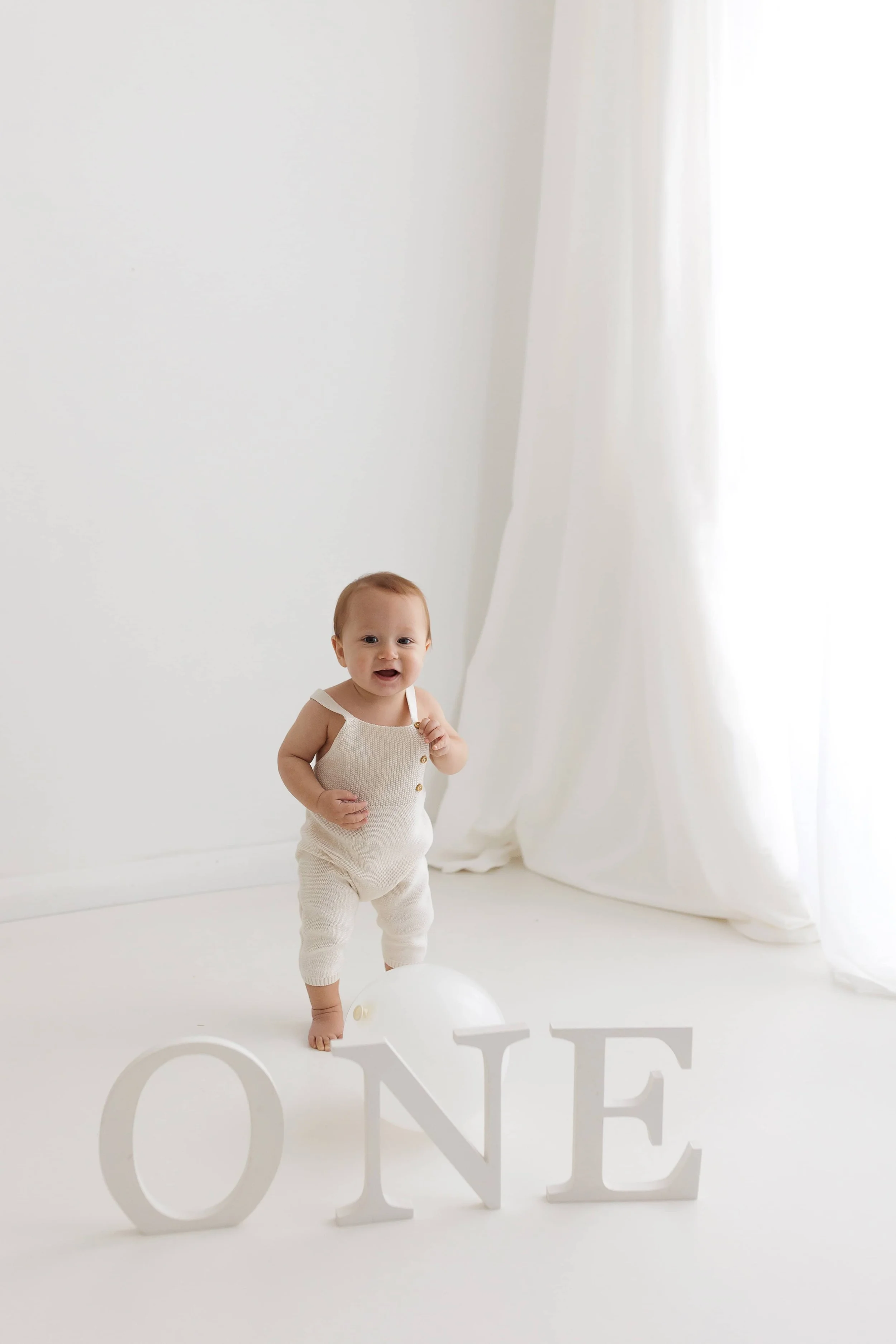 A baby standing on a white floor, holding a small object, with large white letters spelling 'ONE' in front, and a white balloon behind, in a bright room with white curtains.