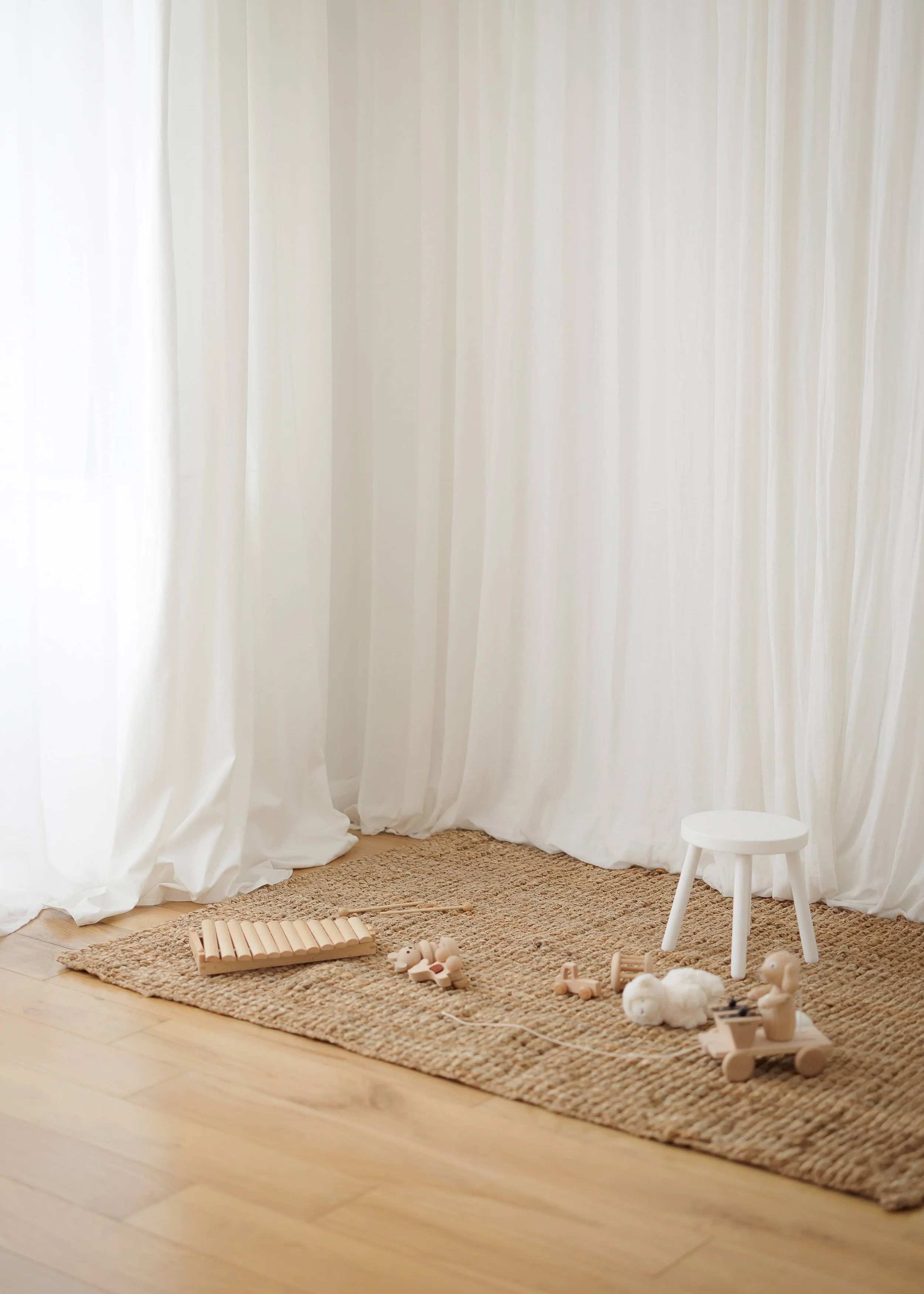 A cozy corner with a textured area rug on wooden floor, decorated with children's toys including a xylophone, wooden animals, and a small white chair, against white curtains.
