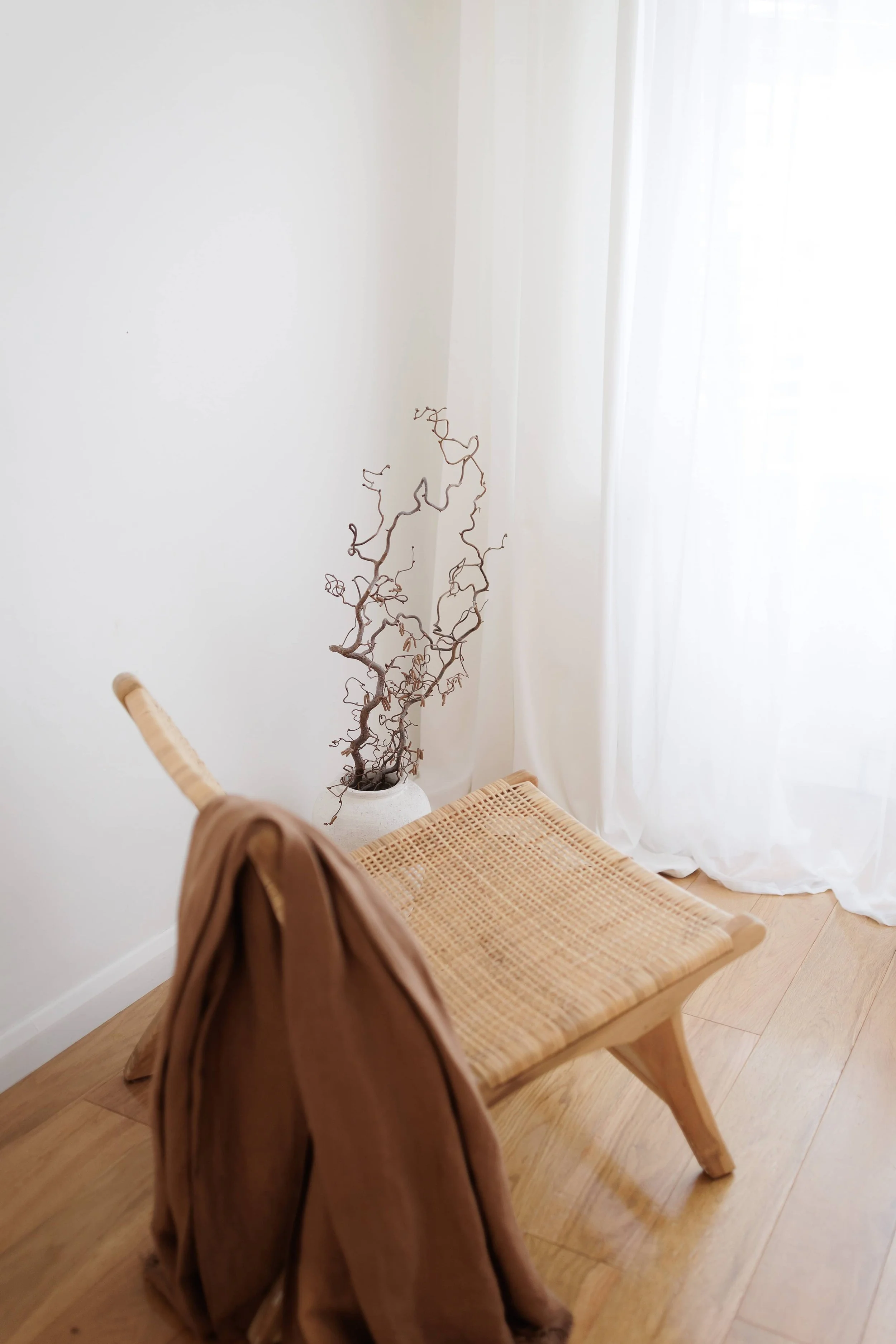 A minimalist room corner with a wooden chair draped with a brown jacket, placed on a hardwood floor, next to a white vase containing curly, leafless branches, and sunlight filtering through sheer white curtains.