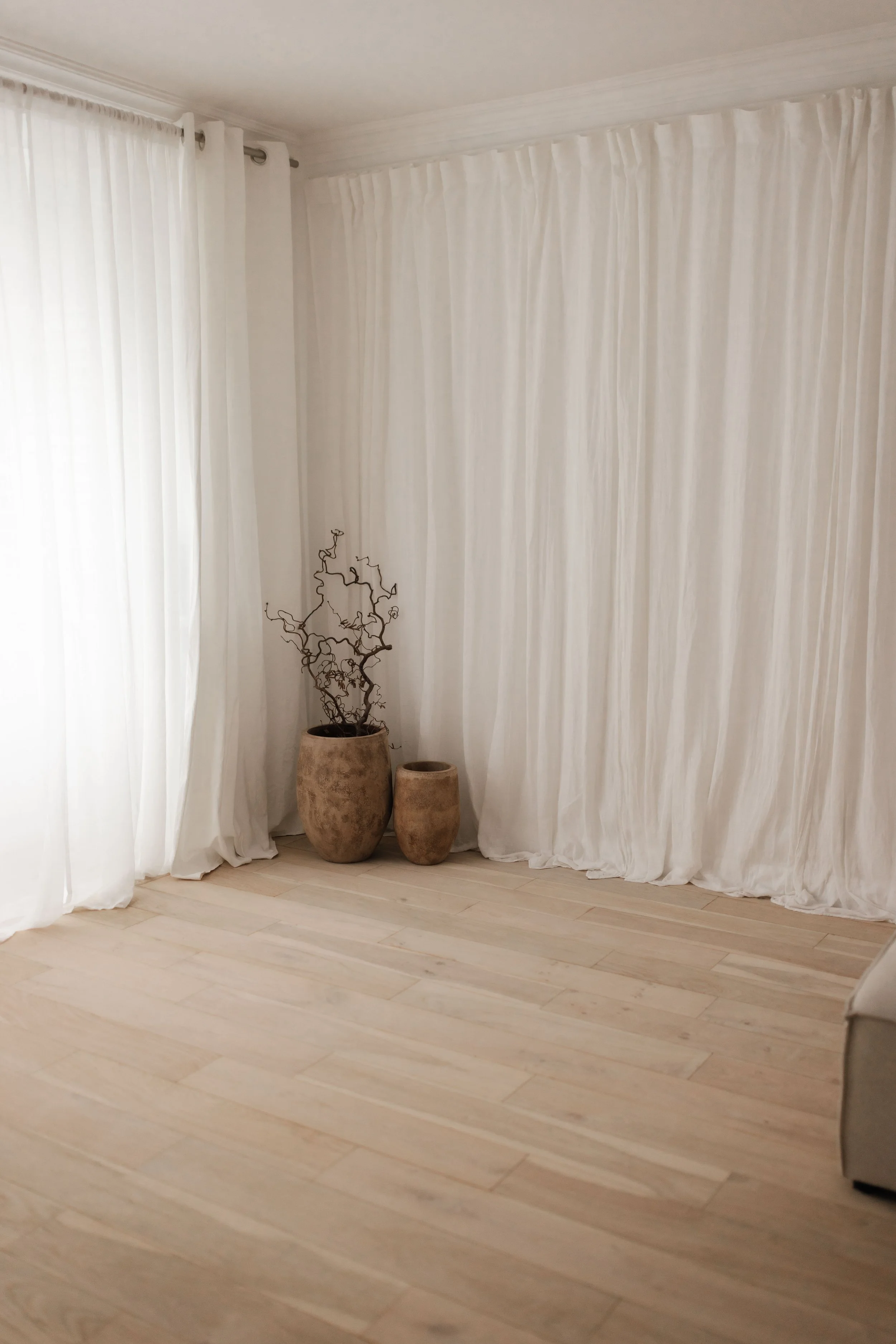 A corner of a photography studio in Surrey with white curtains, a light-colored wooden floor, and three beige pottery vases of different sizes with decorative twigs in the largest vase.