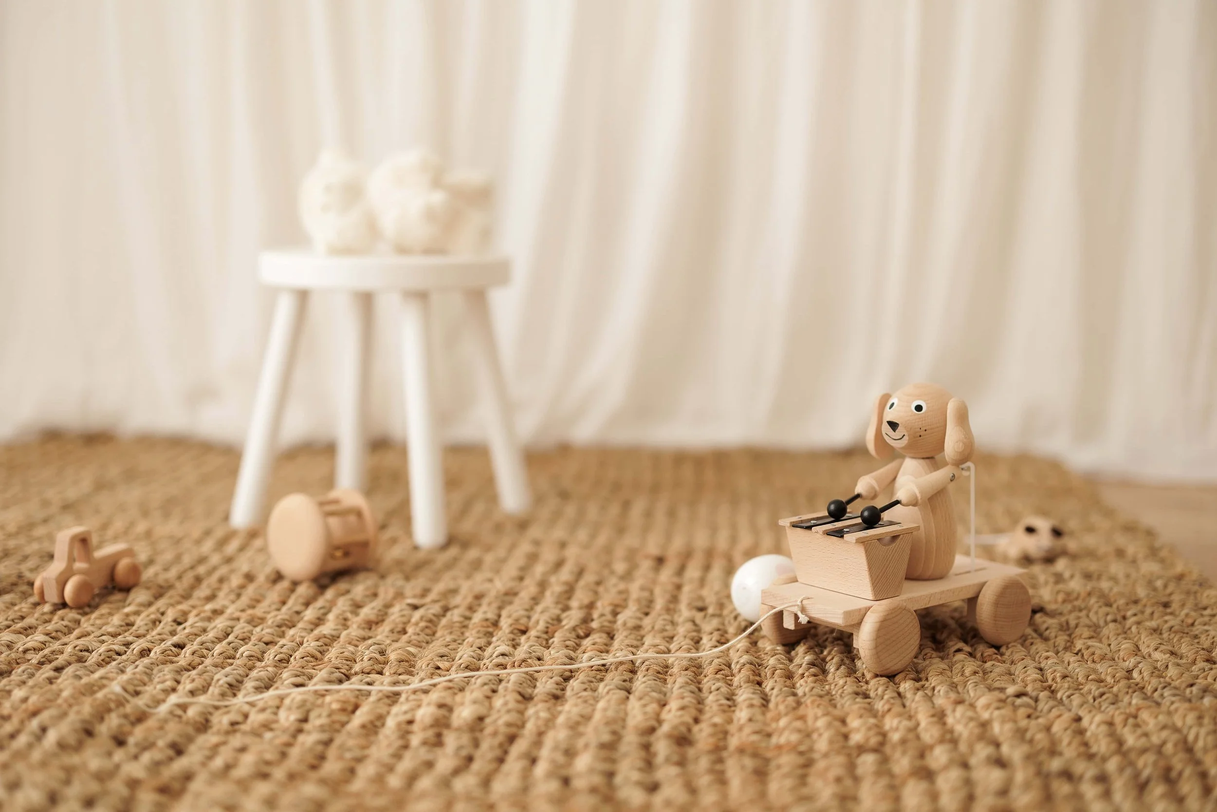 Wooden dog toy riding a cart with a stick to make it move, placed on a woven mat with other small wooden toys and a white stool with plush toys in the background.