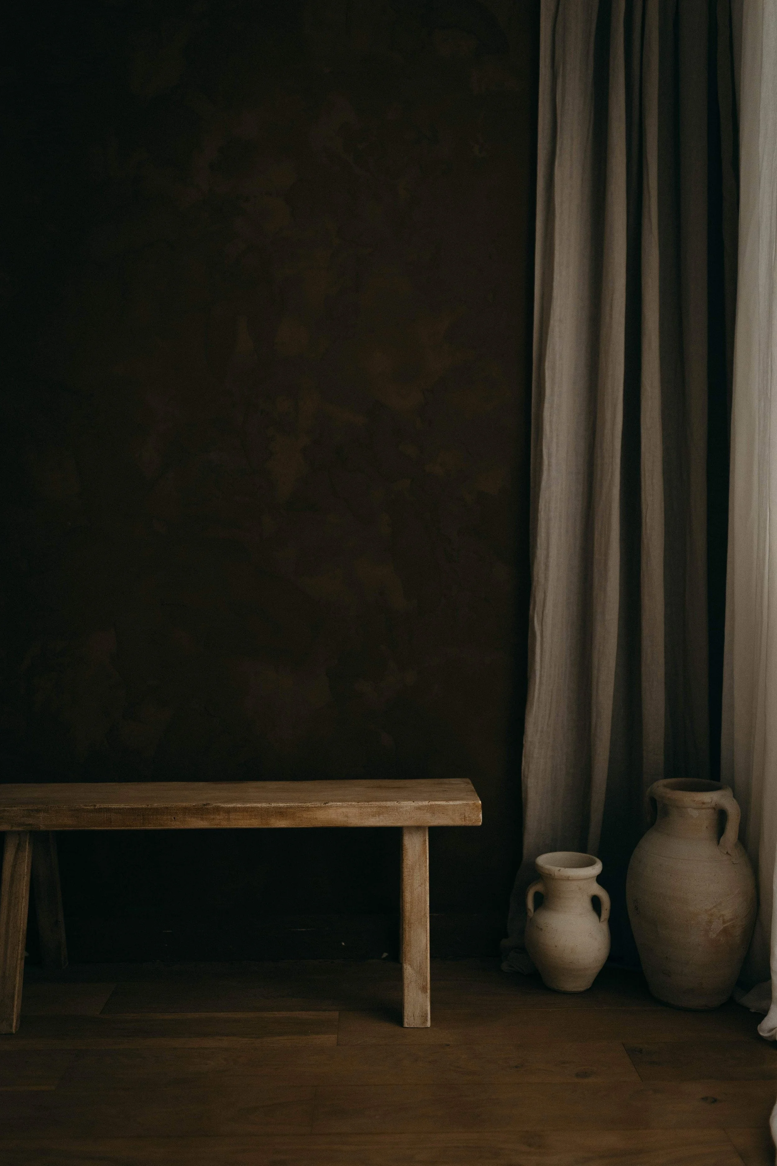 Minimalist interior scene with a light wood bench, two large white pottery vases, dark textured wall, beige curtains on the right side, and wooden floor.
