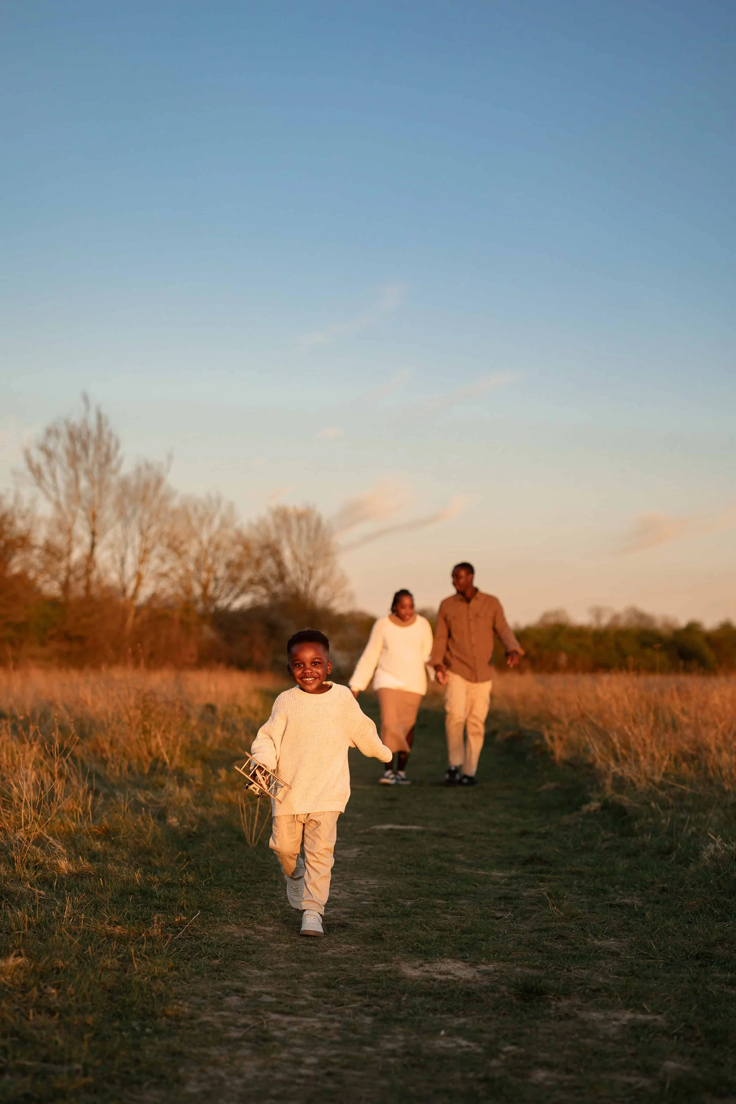 A young child smiling and running on a grassy path with a toy airplane in hand, followed by two adults walking together in the background during sunset in a rural landscape.