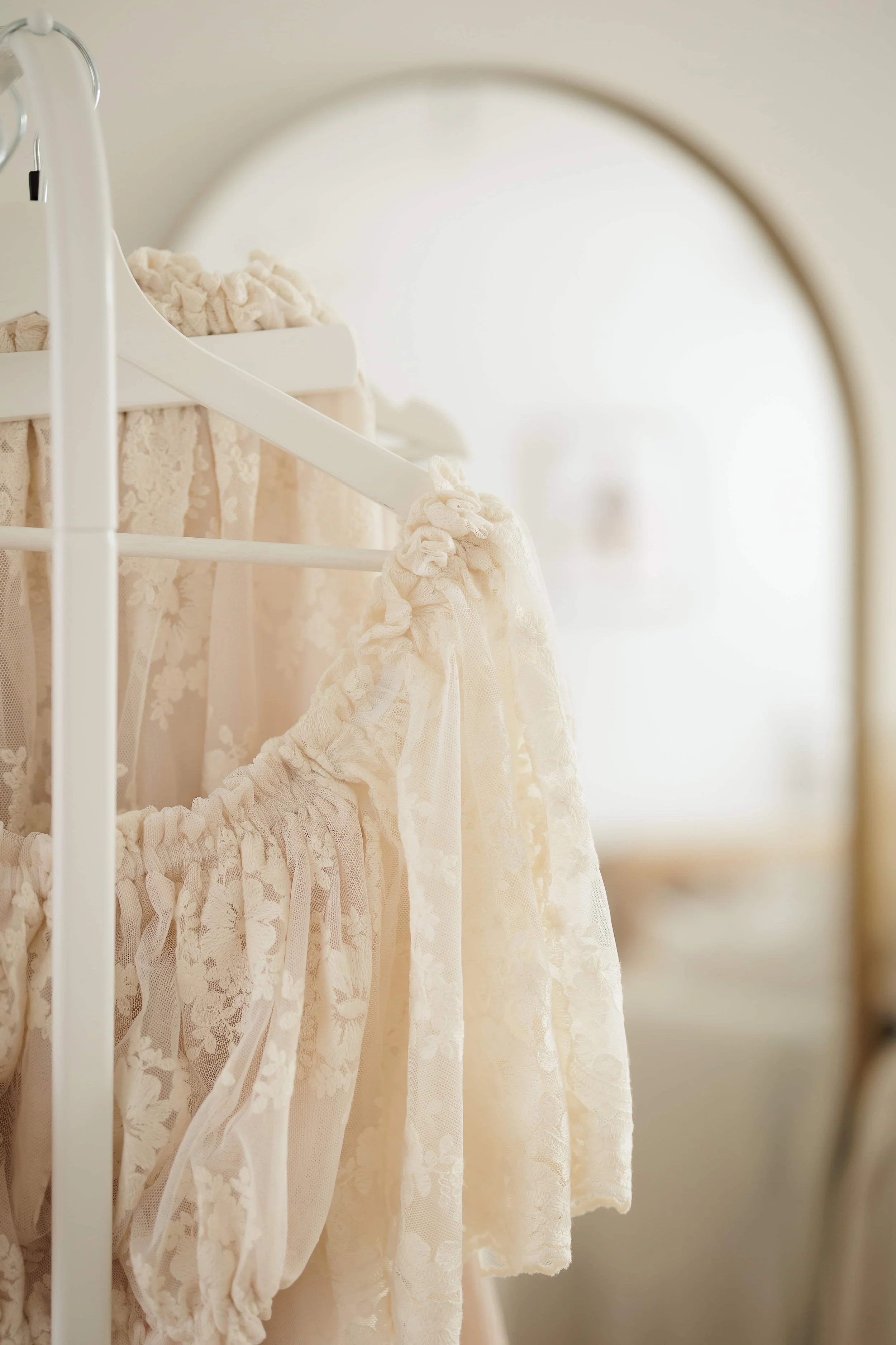 Close-up of delicate, cream-colored lace dresses hanging on a white clothing rack, reflected in an arched mirror.