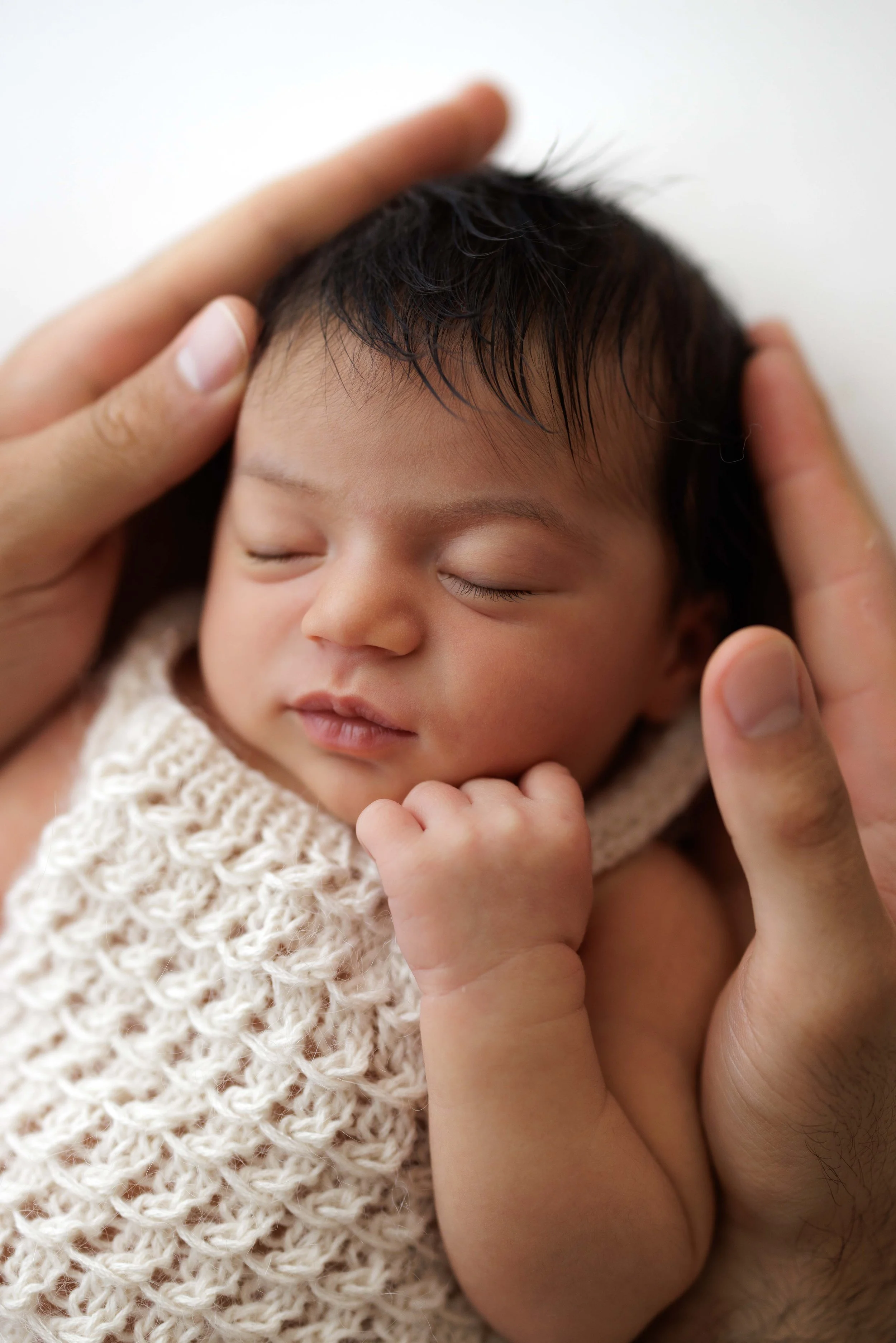 Close-up of a sleeping baby with dark hair, wearing a knitted cream romper, being gently held by an adult hand.