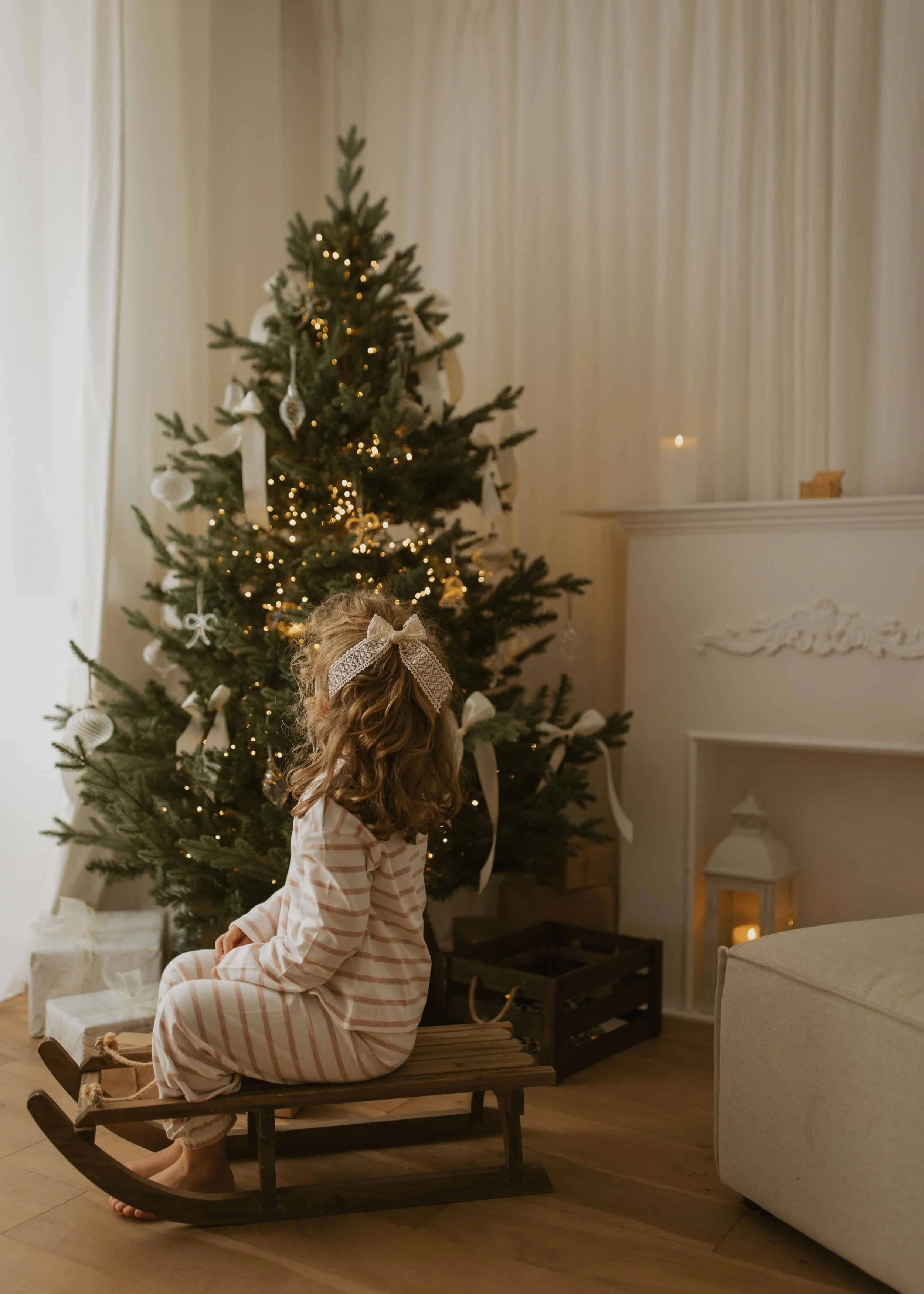 little girl in a pyjama and seated on a wooden sled by a christmas tree decor and a fireplace