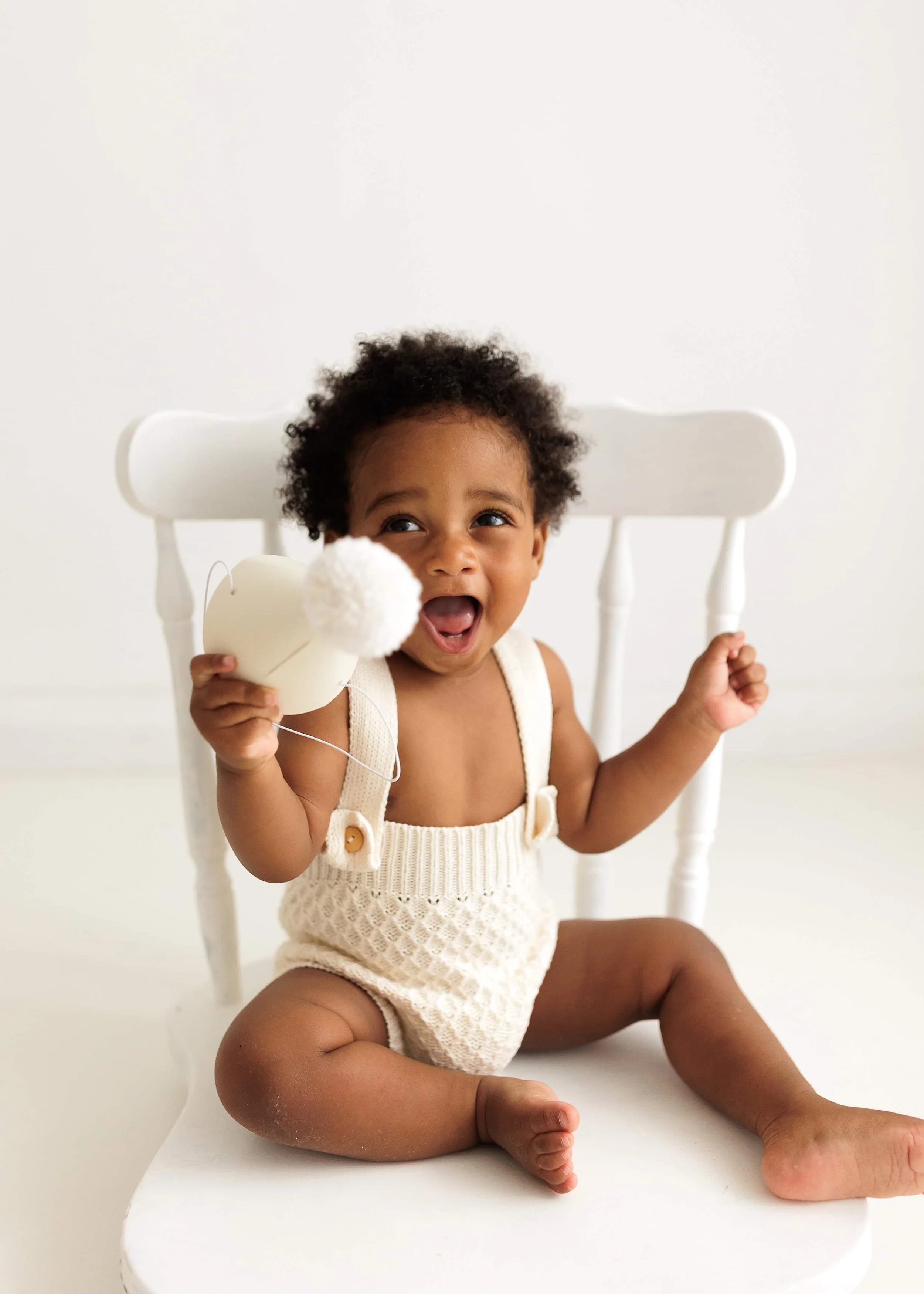 Smiling toddler boy with curly hair sitting on a white chair, holding a white plush toy with a pom-pom, wearing a cream-colored knitted romper, in a bright room.