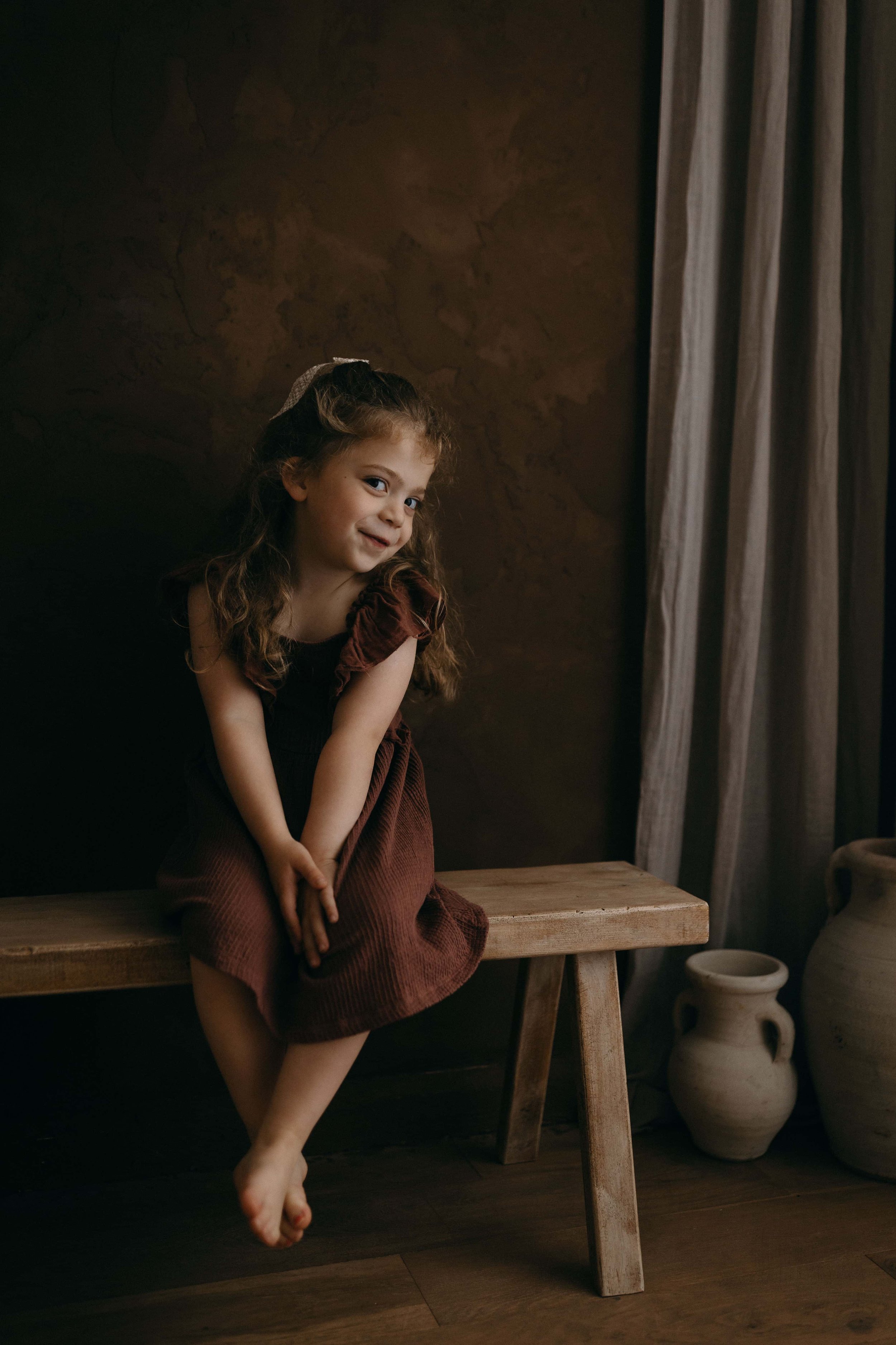 A young girl with curly hair sitting on a wooden bench, smiling, in a room with dark walls, a curtain, and pottery on the floor.