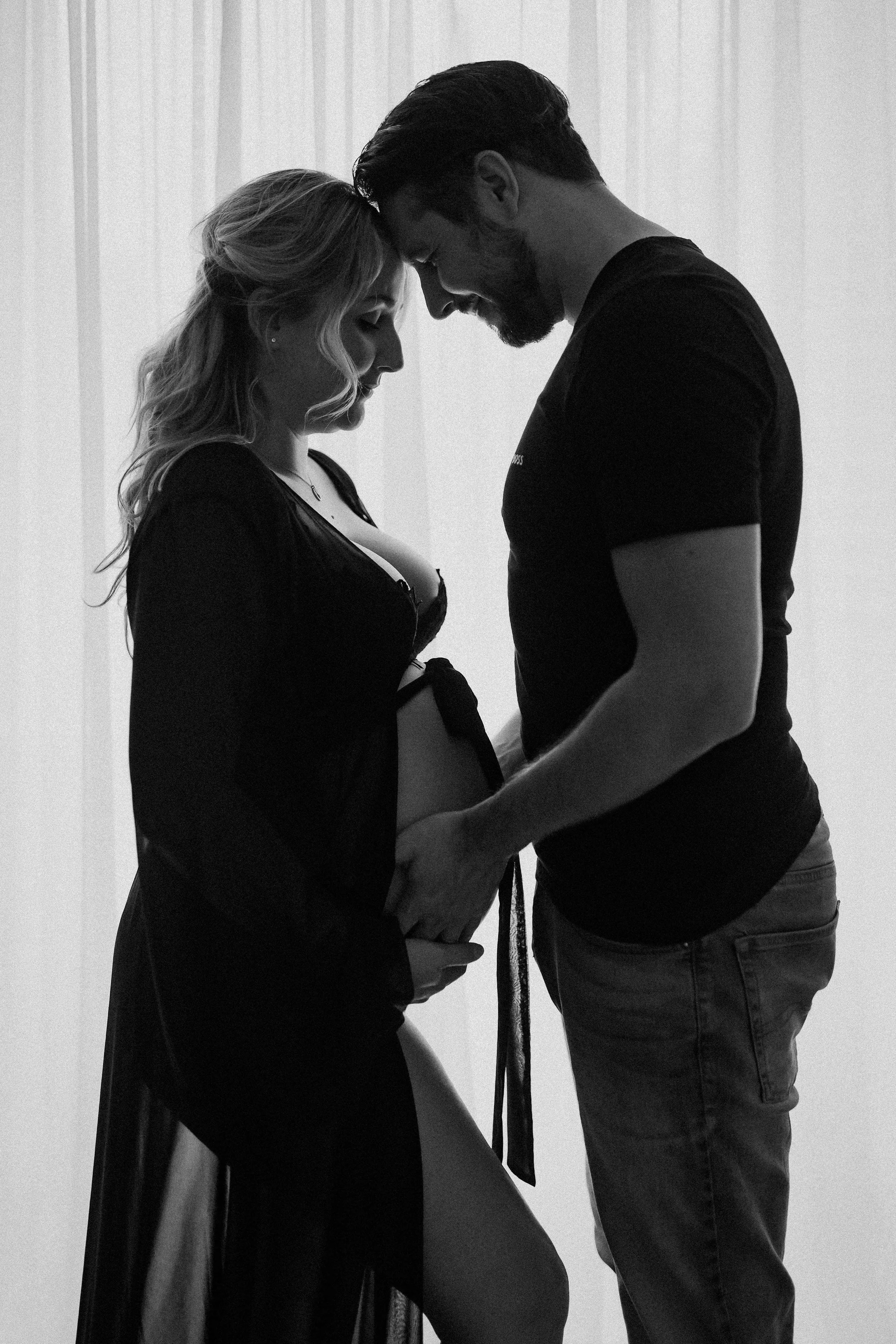 A pregnant woman and her partner touching foreheads and holding hands, with the woman wearing a black dress and the man in a black t-shirt.