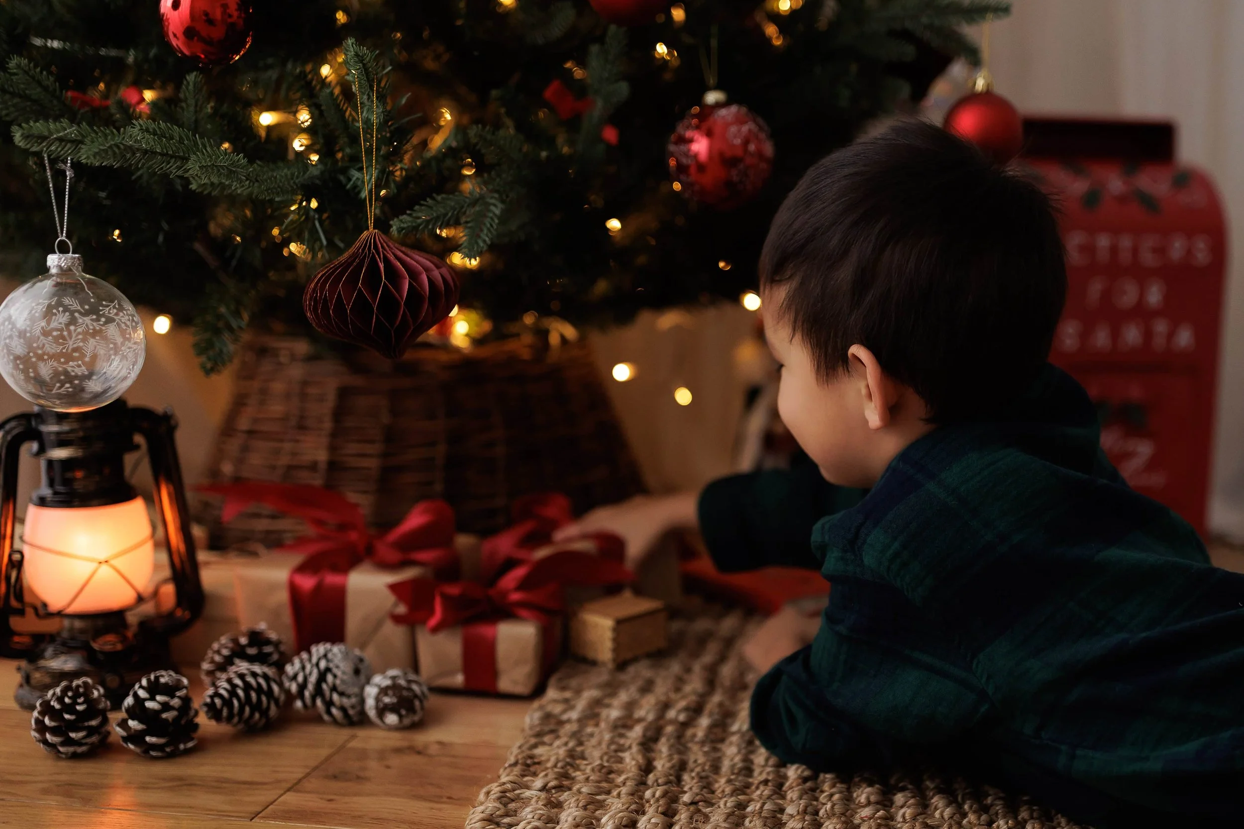 A young boy with dark hair lying on the floor in front of a decorated Christmas tree, surrounded by wrapped presents, pinecones, and a lantern, with soft warm lighting and Christmas ornaments.