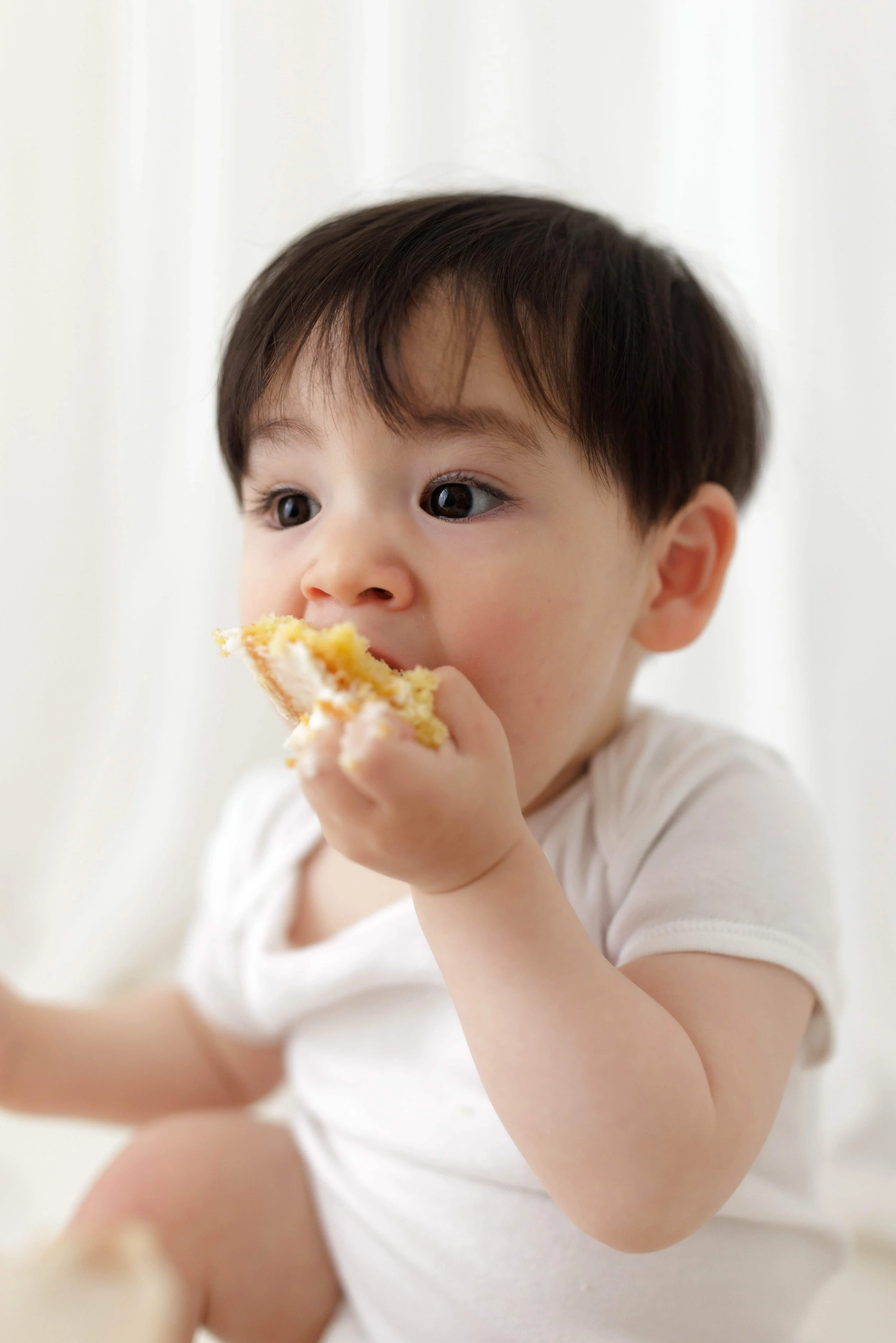 A one year old baby boy with dark hair and big eyes eating a piece of cake for his smash cake photoshoot