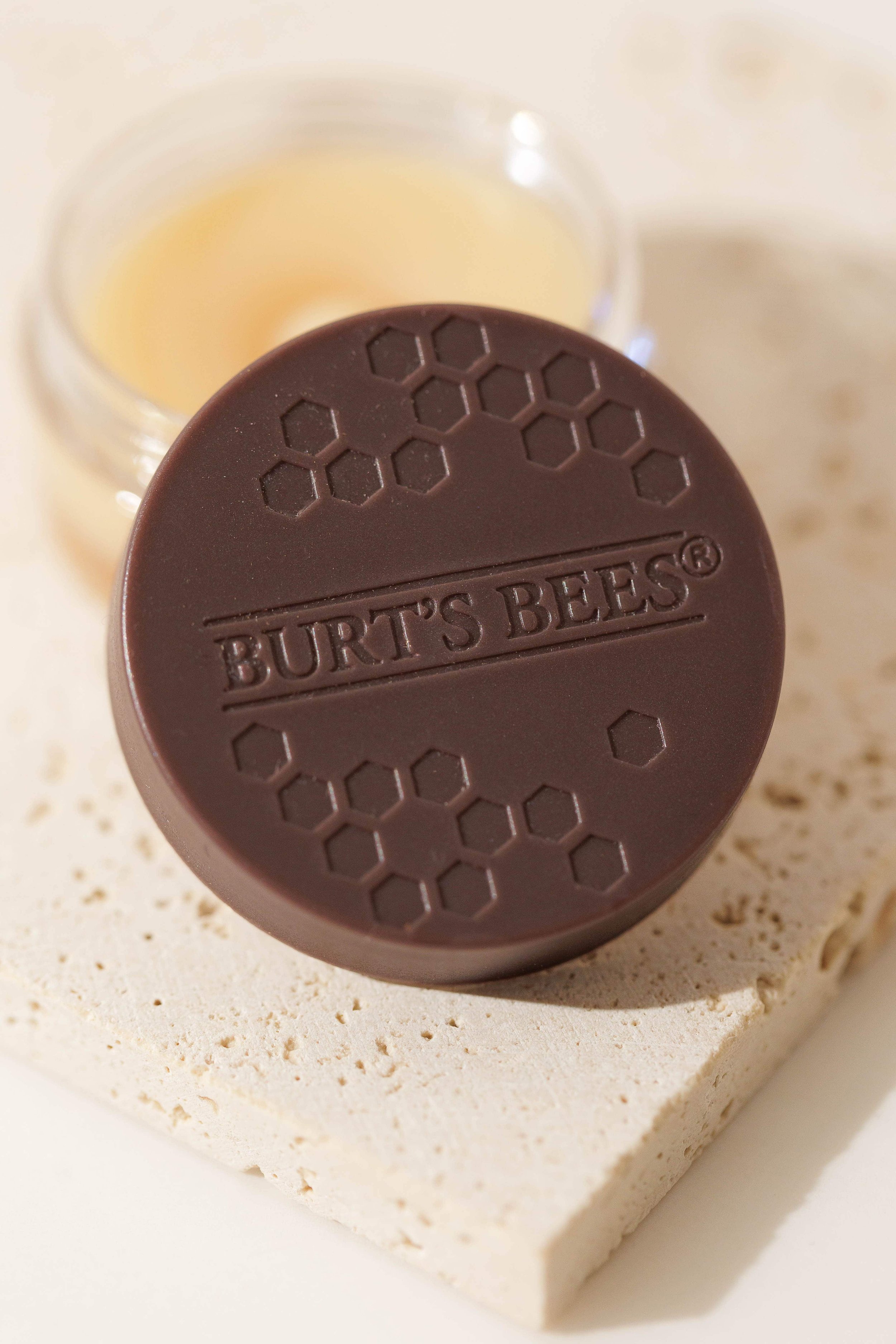 Close-up of a jar of burt bee lip mask with honeycomb pattern and 'Burt's Bees' embossed on it, resting on a white textured surface 