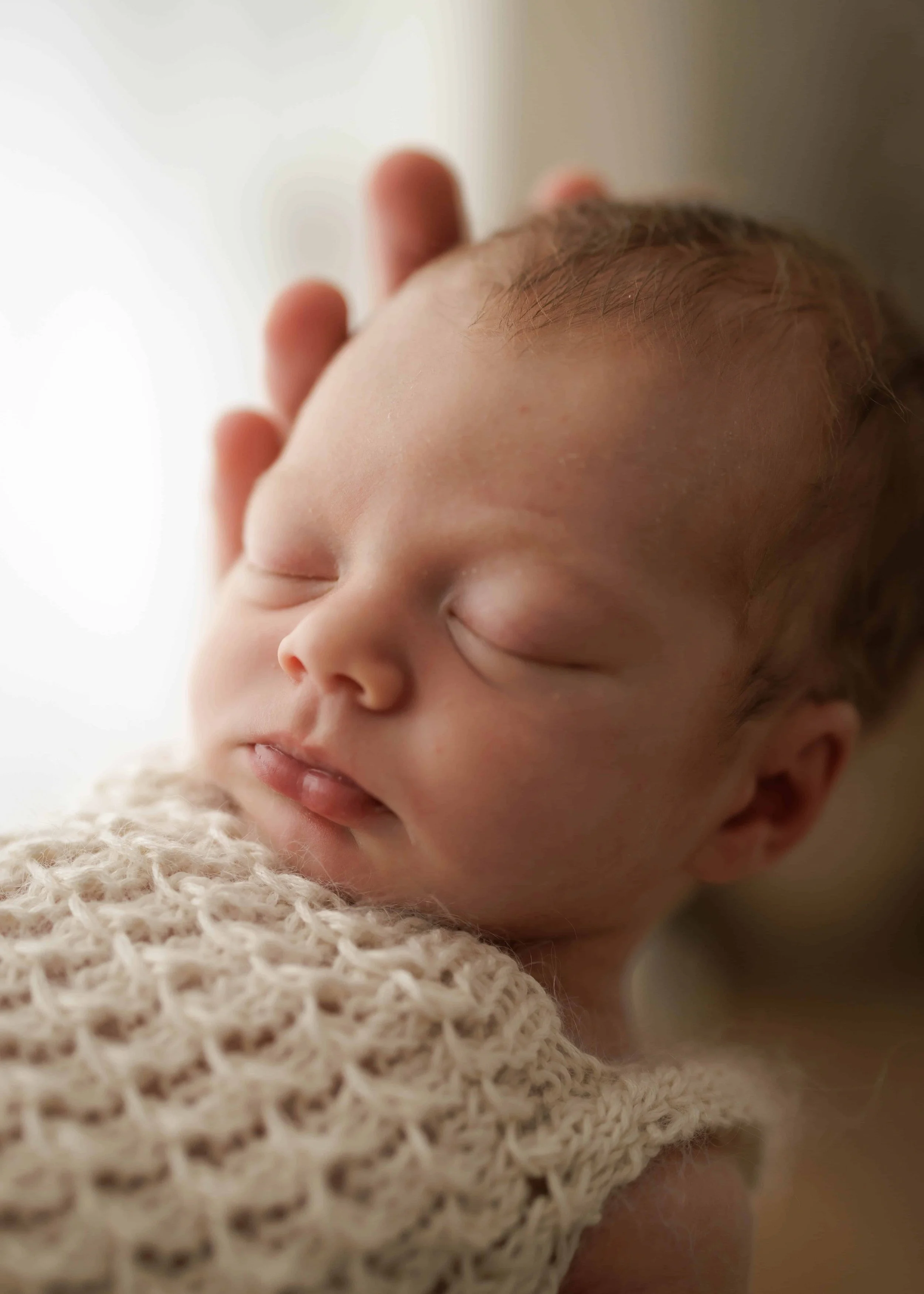 Close-up of a sleeping newborn baby with a hand gently touching its head, lying on a textured cream-colored blanket.