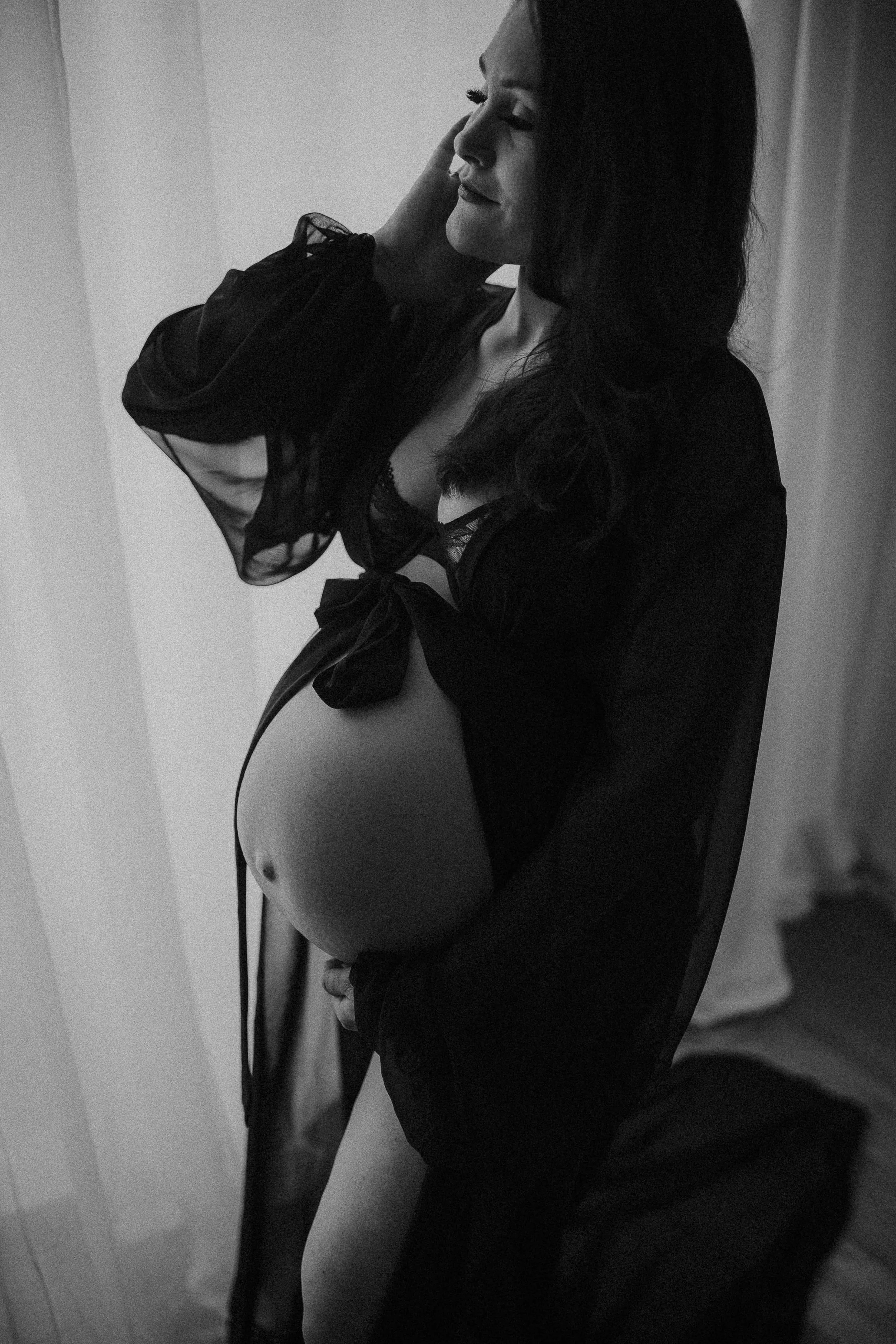Black and white photo of a pregnant woman in lingerie and a sheer robe, smiling with her eyes closed, touching her hair and belly.