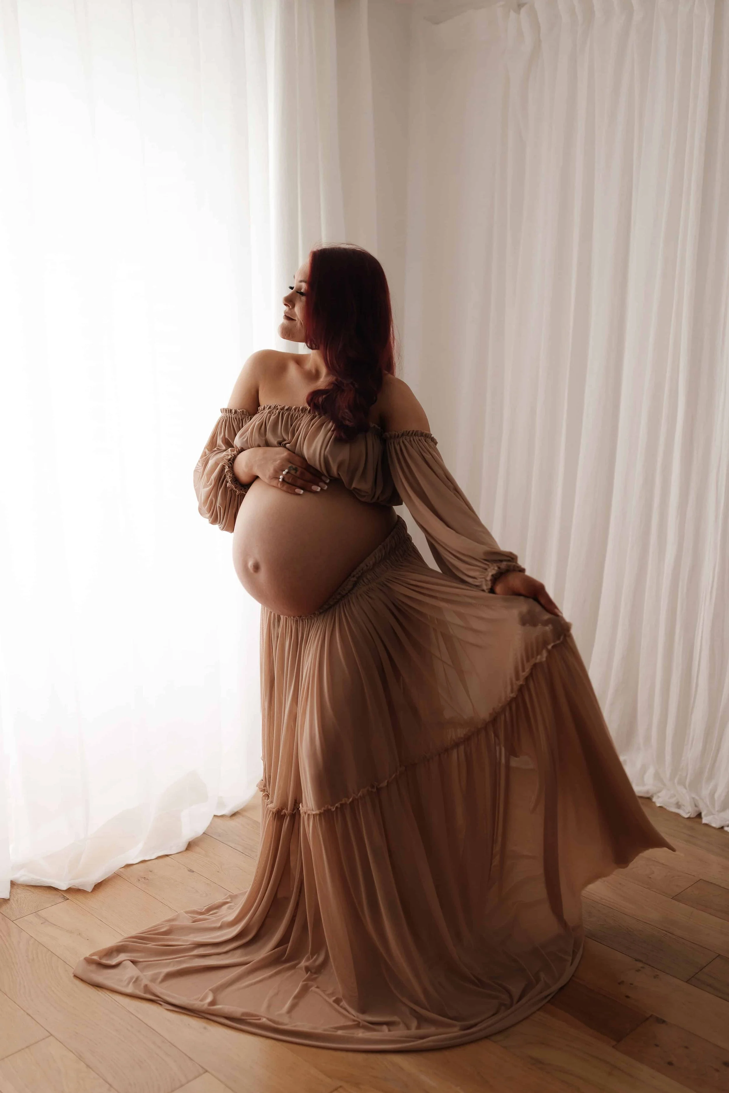Pregnant woman in beige flowing dress standing near window with sheer curtains, touching her belly, in a softly lit room.