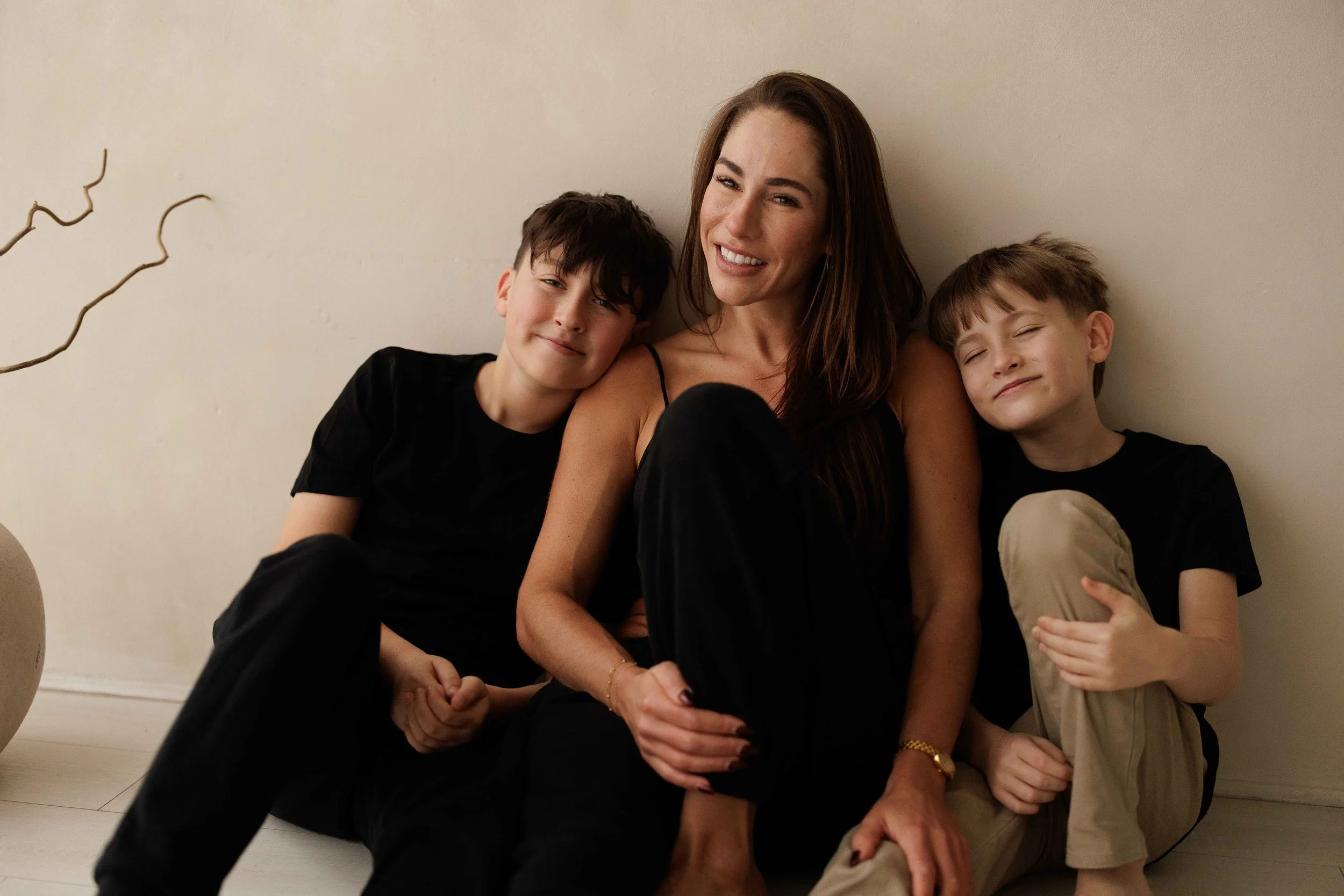 A mum sitting on the floor with her two young boys, all smiling and leaning against a light-colored wall.