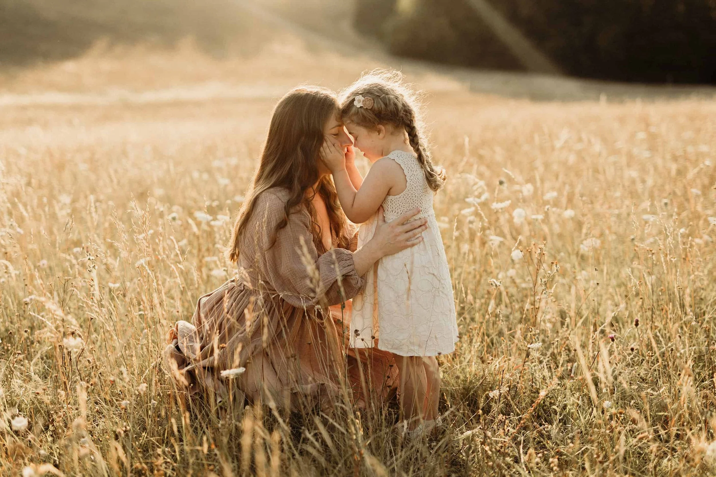 A woman and a young girl touching foreheads and smiling in a field of tall grass during sunset.