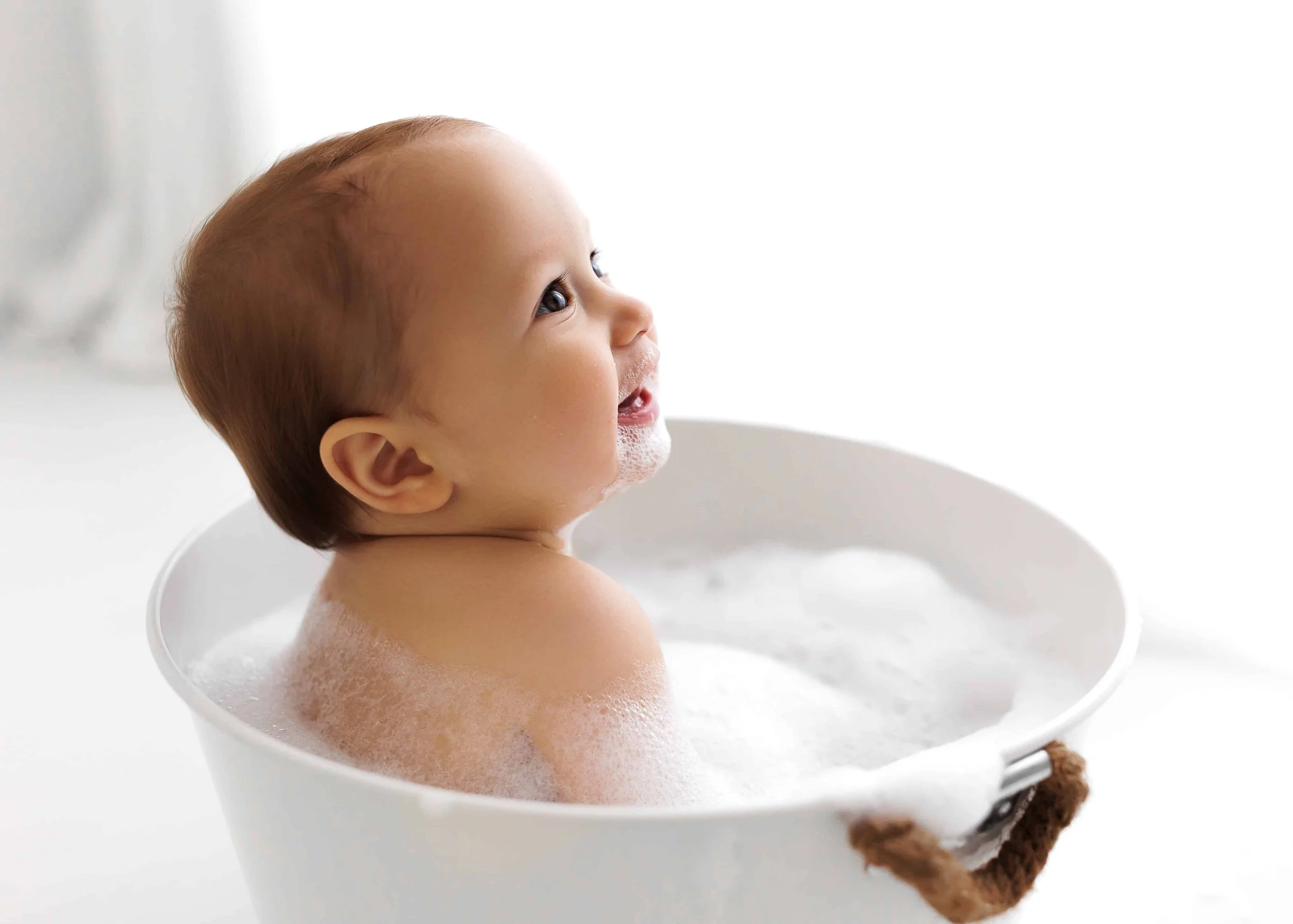 Young child with light brown hair enjoying a bath in a white tub with soap bubbles.