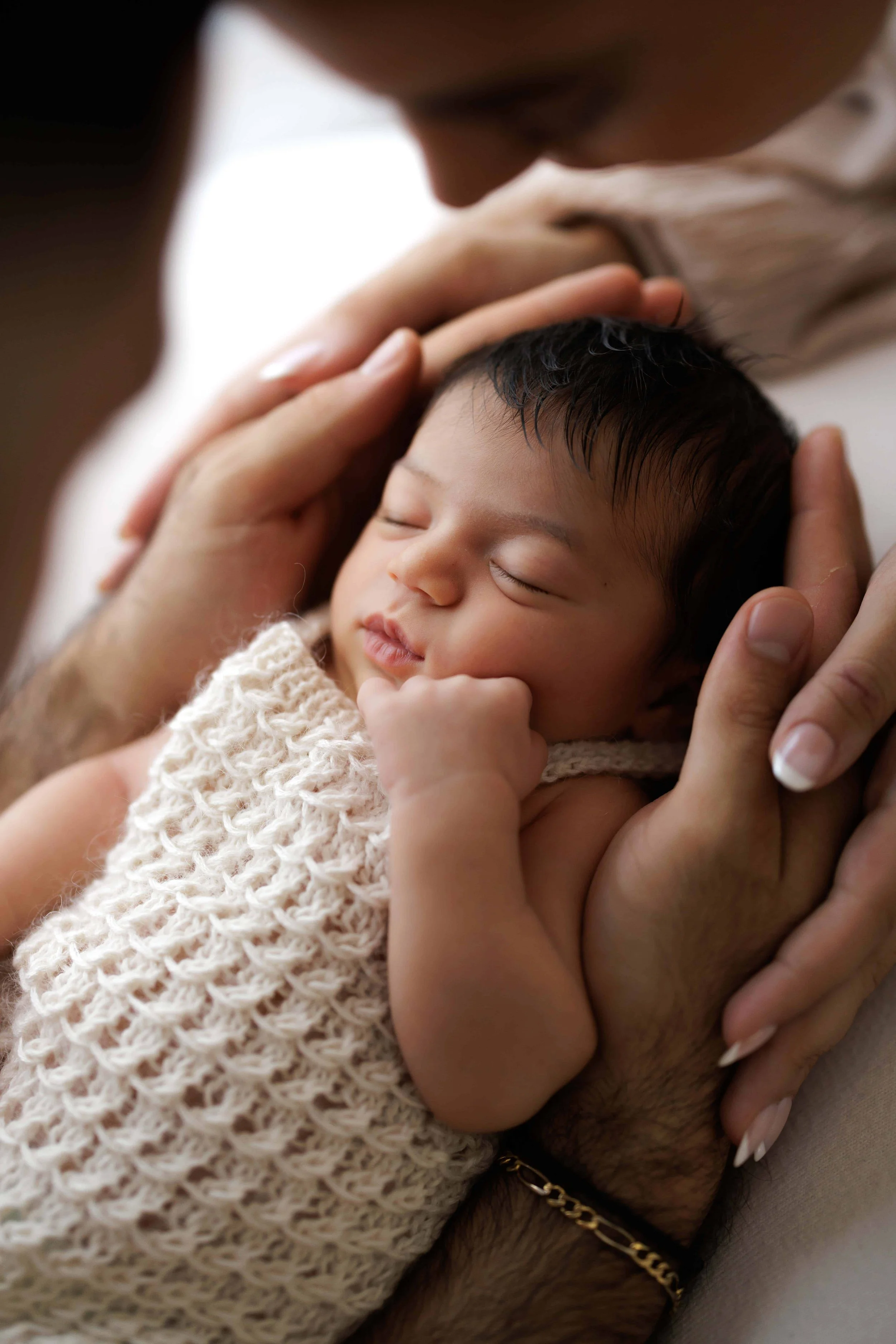 Close-up of a person holding a sleeping baby with dark hair, wearing a knitted cream-colored garment, with a gentle expression.