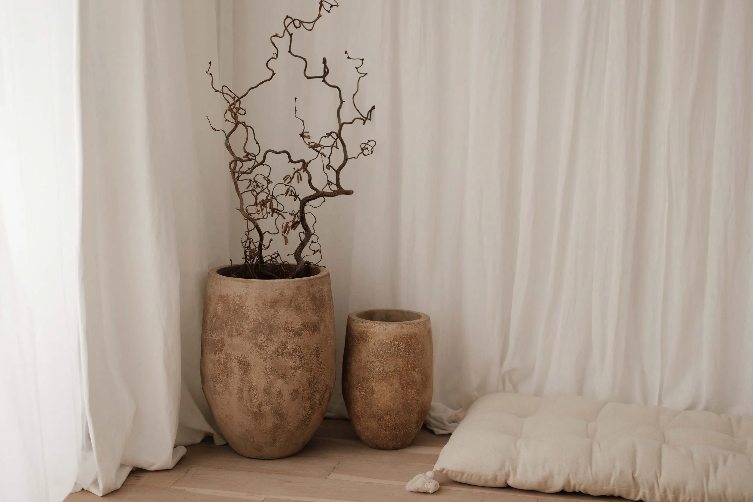 A minimalistic mediterranean studio with two large clay pots, one containing a leafless, twisted, dry plant, placed on a light wooden floor against white curtains, along with a cushioned sleeping pad on the floor.