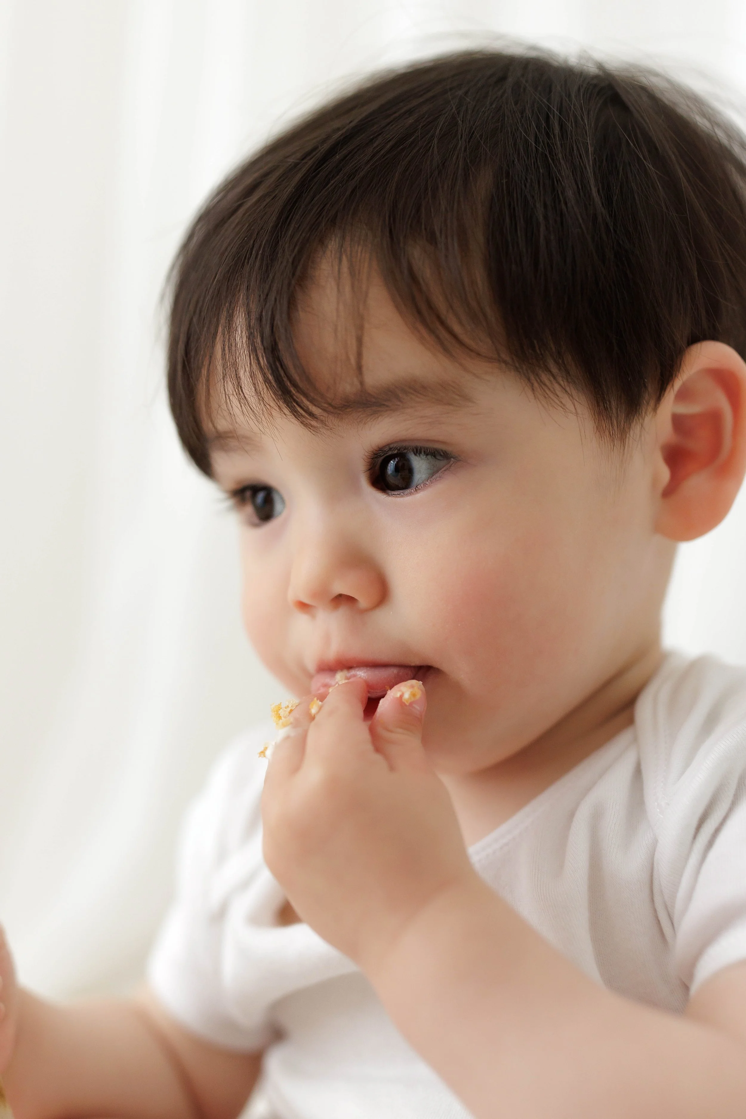 Close-up of a baby boy with dark hair and brown eyes eating with fingers, wearing a white shirt.
