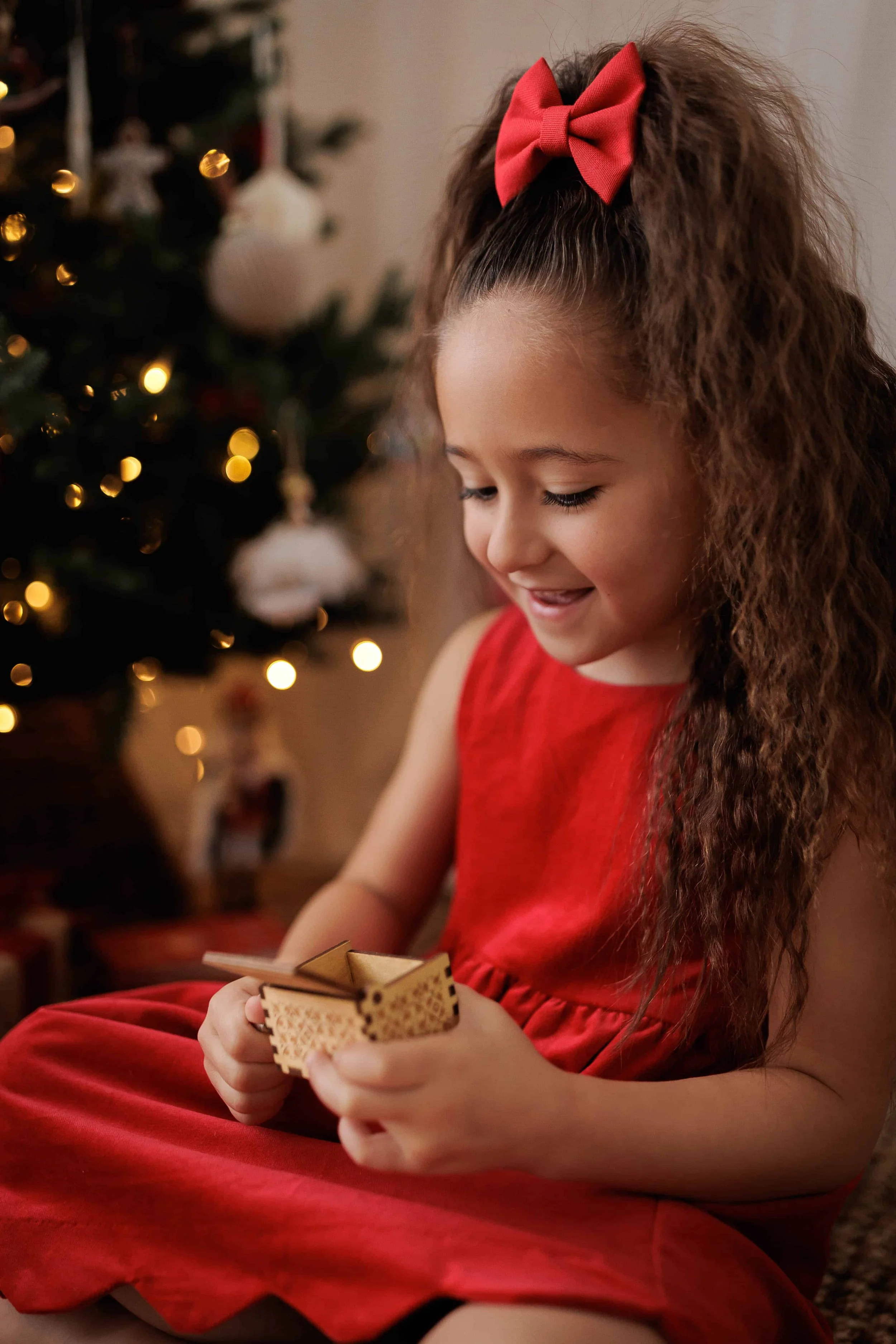 A young girl with curly hair, wearing a red dress and a matching red bow, sitting indoors during Christmas, opening a small gift with a happy expression, with a decorated Christmas tree in the background.