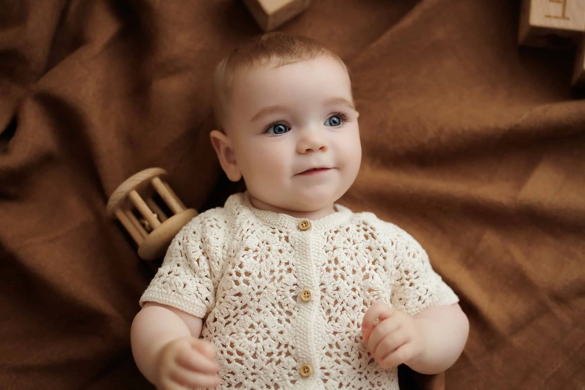 A young baby with blue eyes and light brown hair lying on a brown blanket, wearing a cream crocheted sweater with wooden buttons. A wooden rattle rests near the baby's head.