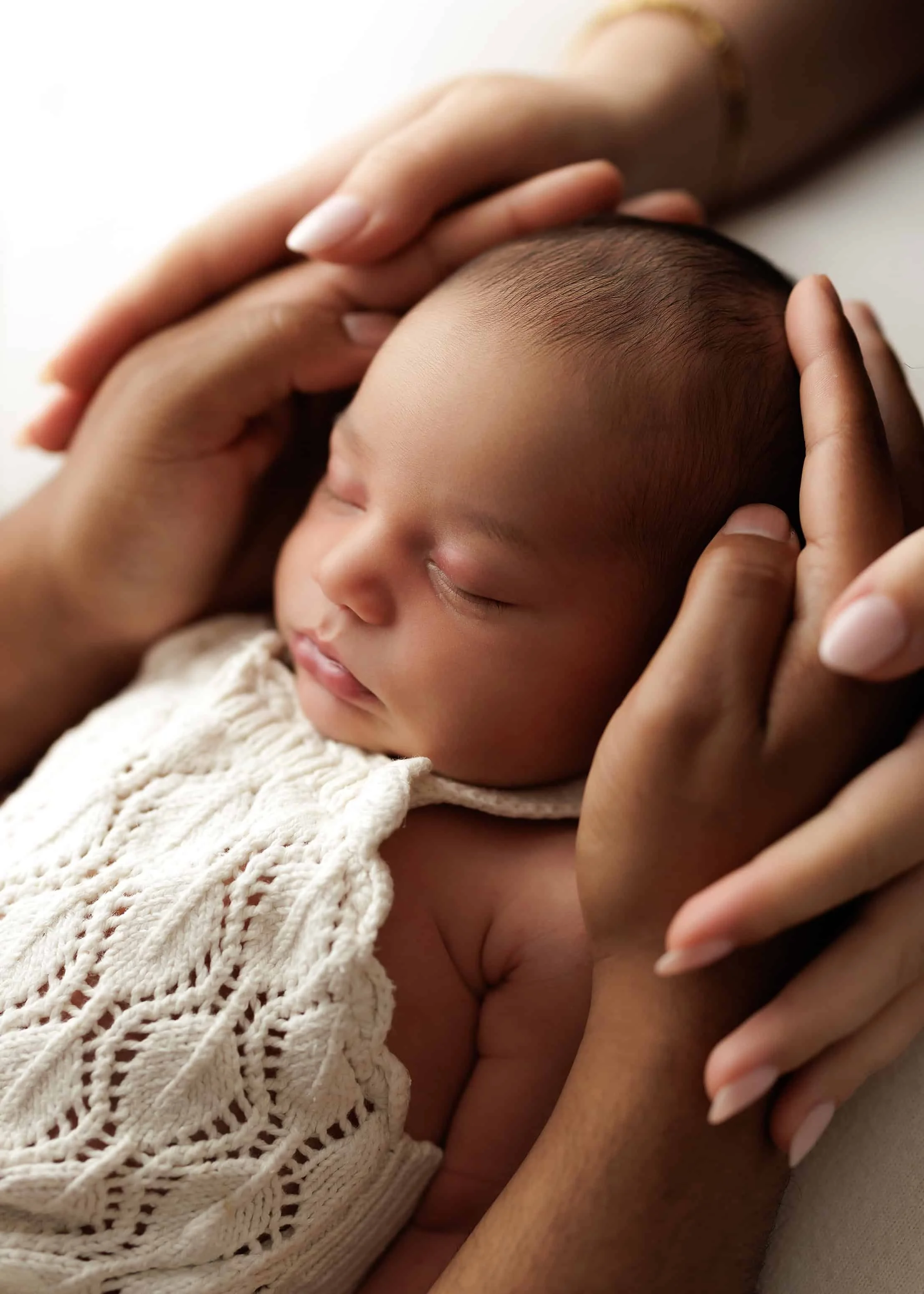 Close-up of a sleeping baby with hands gently touching their head, wearing a cream-colored knit sweater.