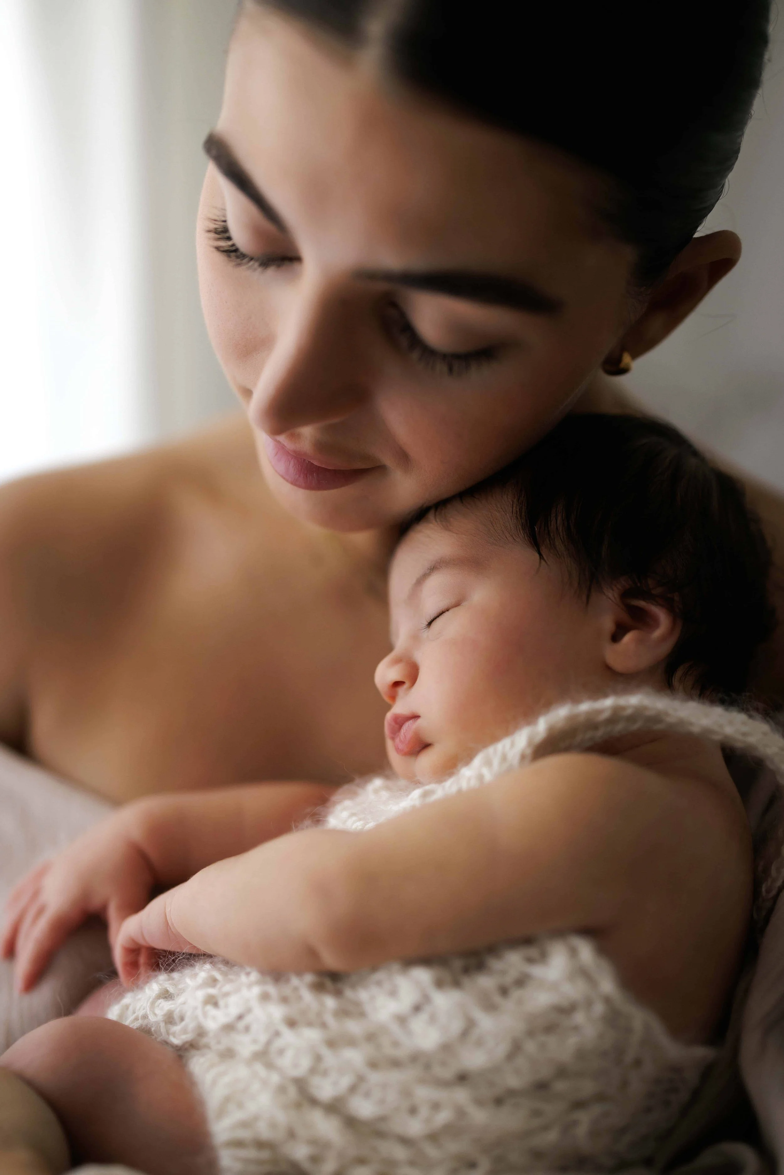 A close-up photograph of a woman with dark hair in a bun holding a sleeping baby wearing a white hand knitted romper