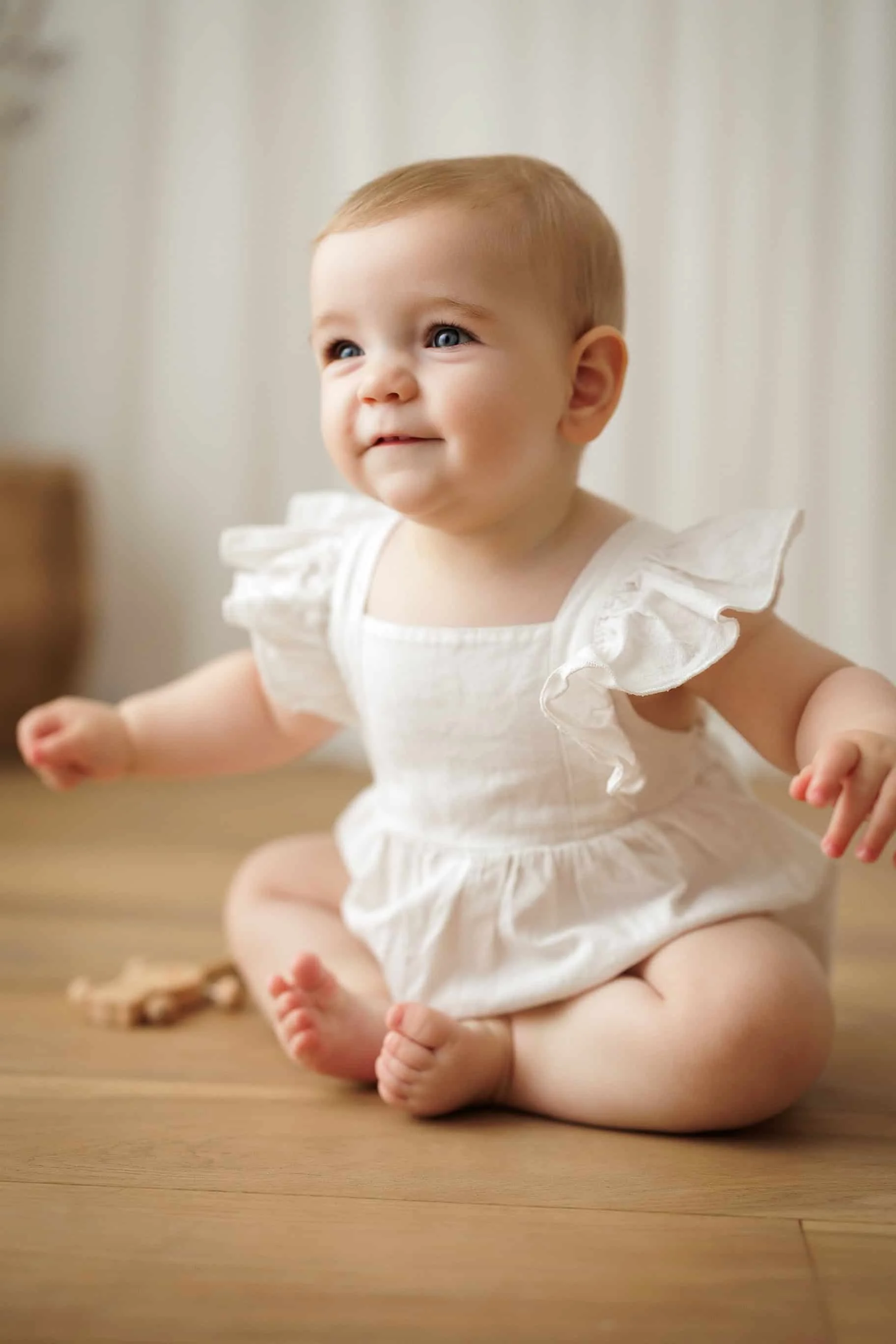 A baby with blonde hair and blue eyes sitting on a wooden floor, wearing a white dress with frilled sleeves.