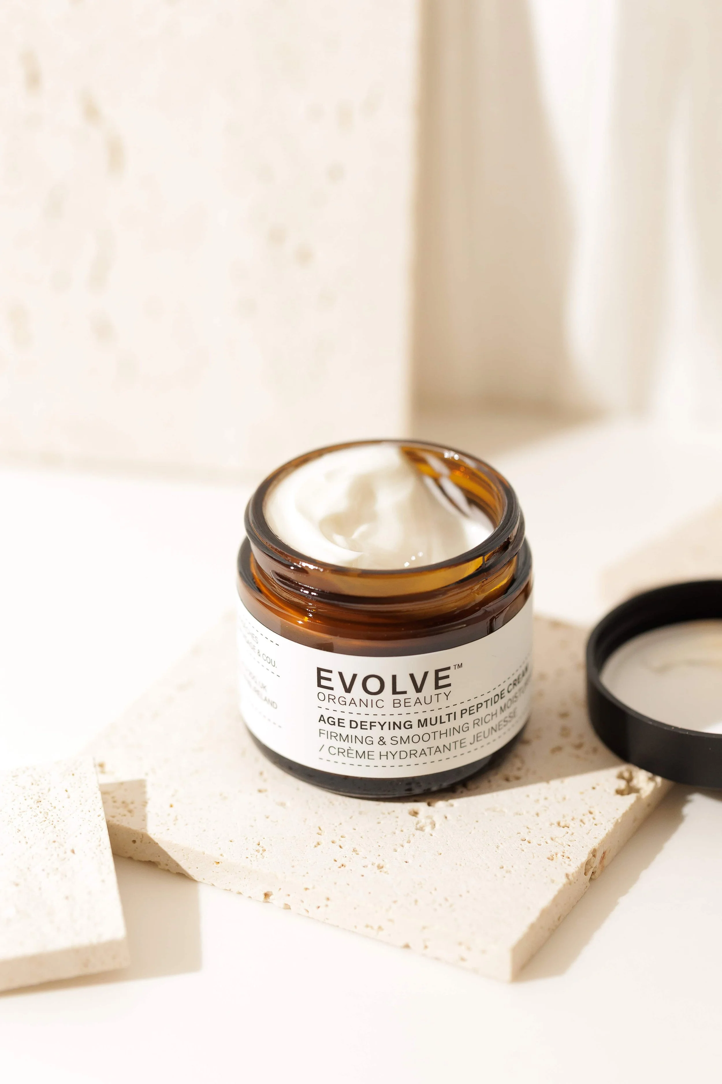 Open jar of EVOLVE organic beauty age defying multi peptide cream with white cream inside, placed on a textured white tile with a black lid beside it, with soft background light.