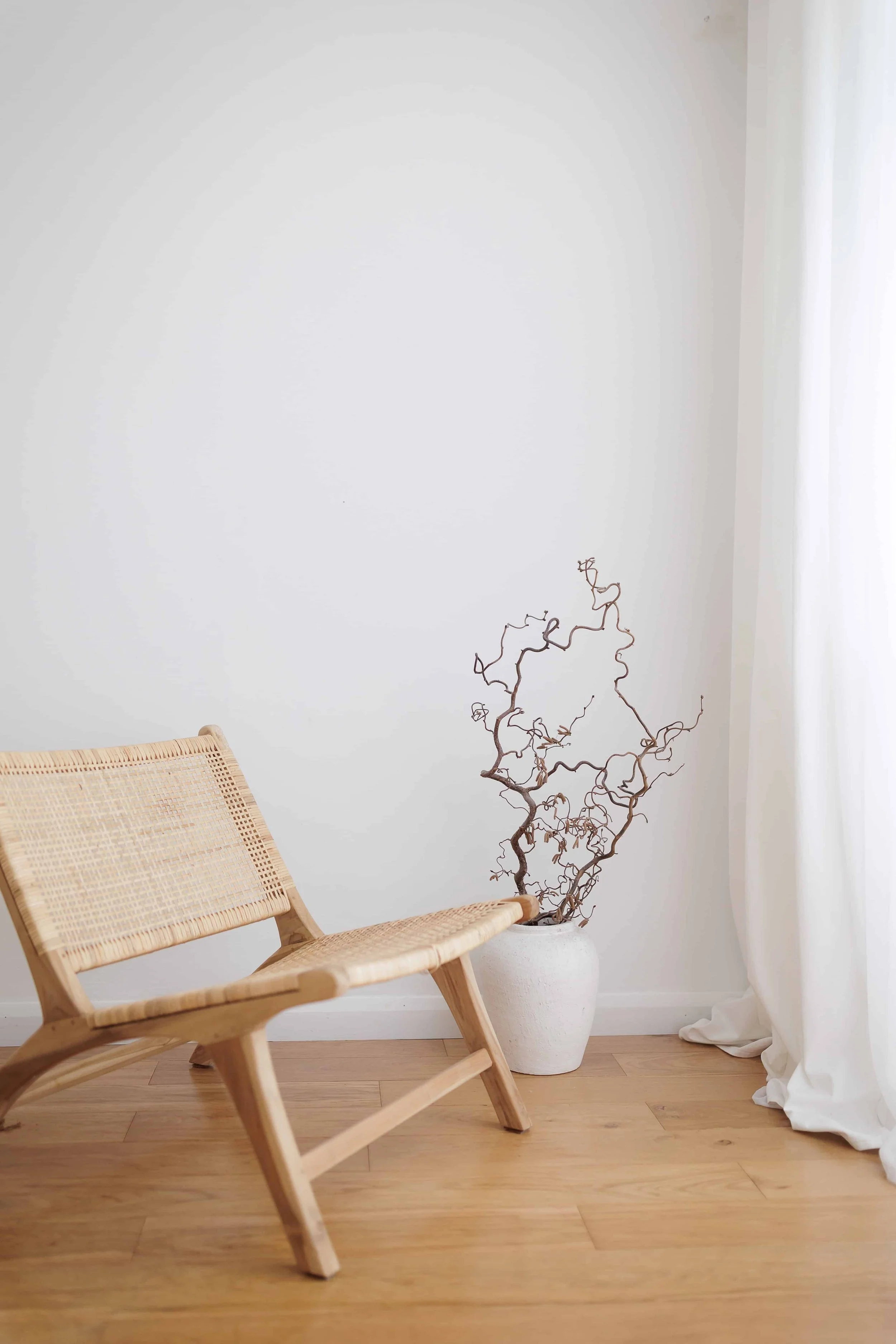 Minimalist interior with a wooden chair, a white vase with dried branches, white curtains, and a wooden floor.