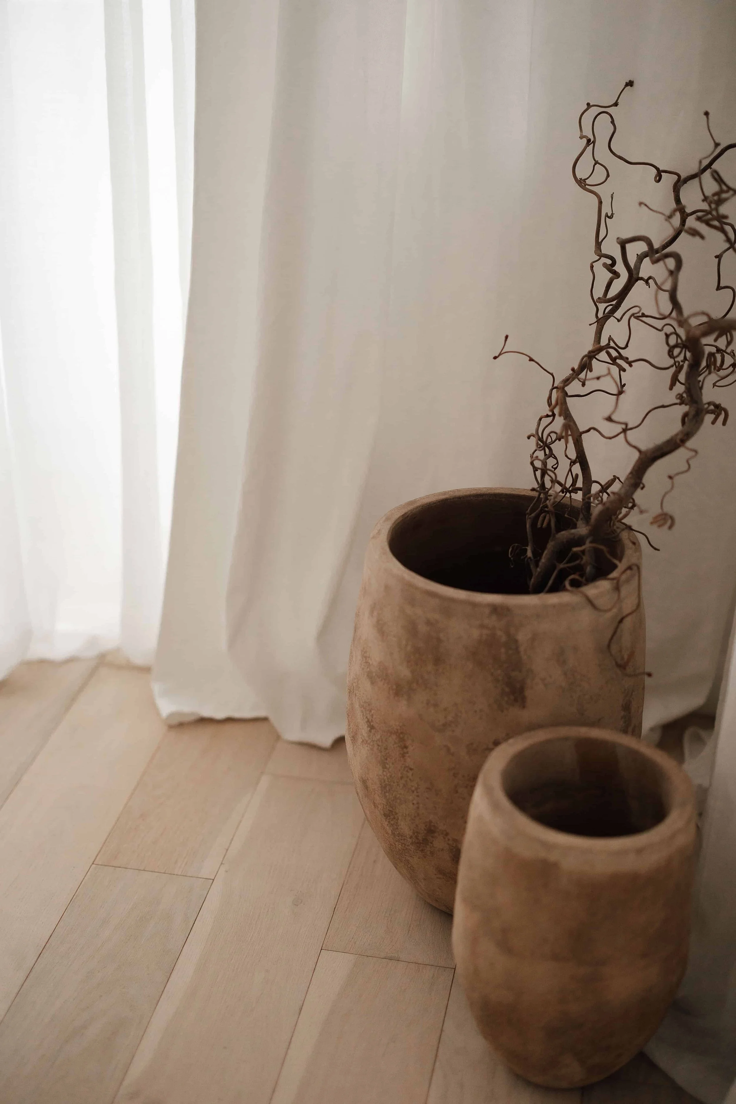 Two large, round, beige pottery planters with dried, twisted twigs inside, placed on a light wood floor near white curtains.