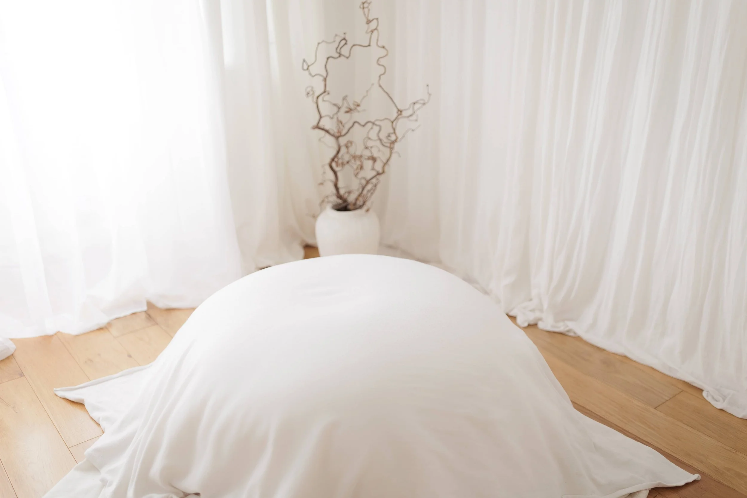 A room with white curtains, a white vase with dried branches in the back, and a white bed or seating area covered with a white sheet or fabric on a wooden floor.