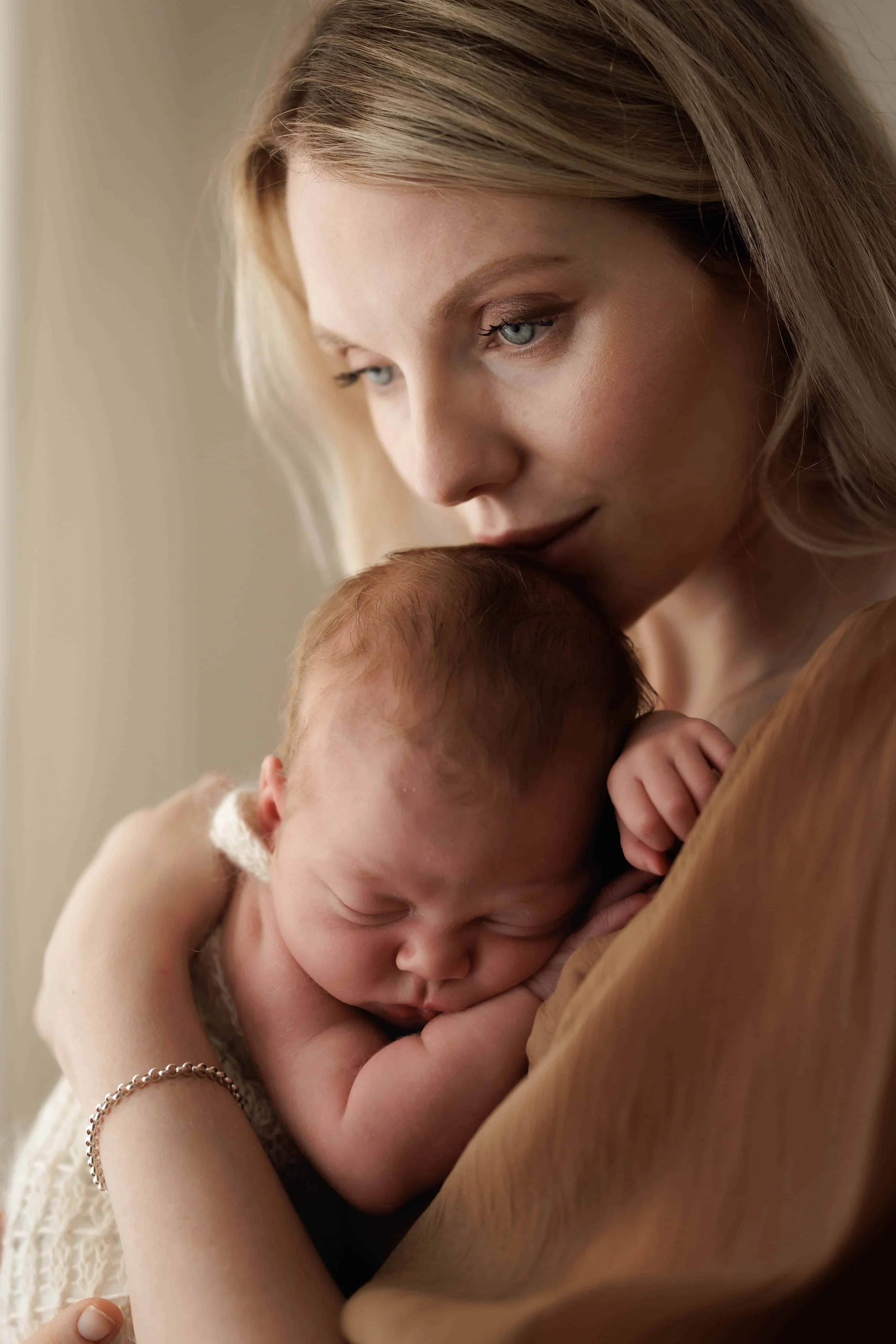 A woman with blonde hair gently kisses the forehead of a sleeping baby with red hair. The scene is tender and intimate, highlighting a moment of love and care.