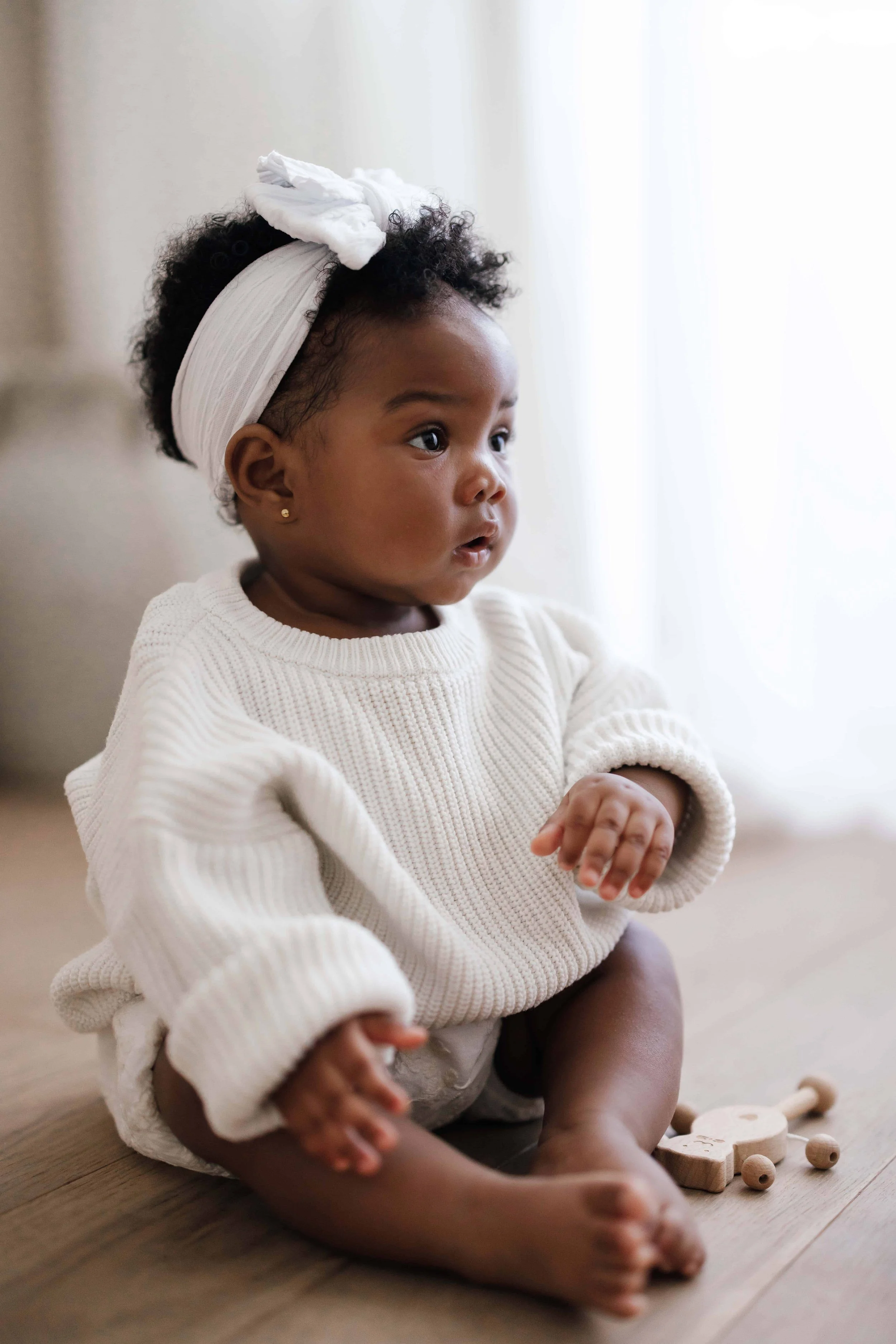 A 9 months old baby girl with curly hair wearing a white headband and sweater, sitting on a wooden floor, with toys nearby, looking attentively to the side.