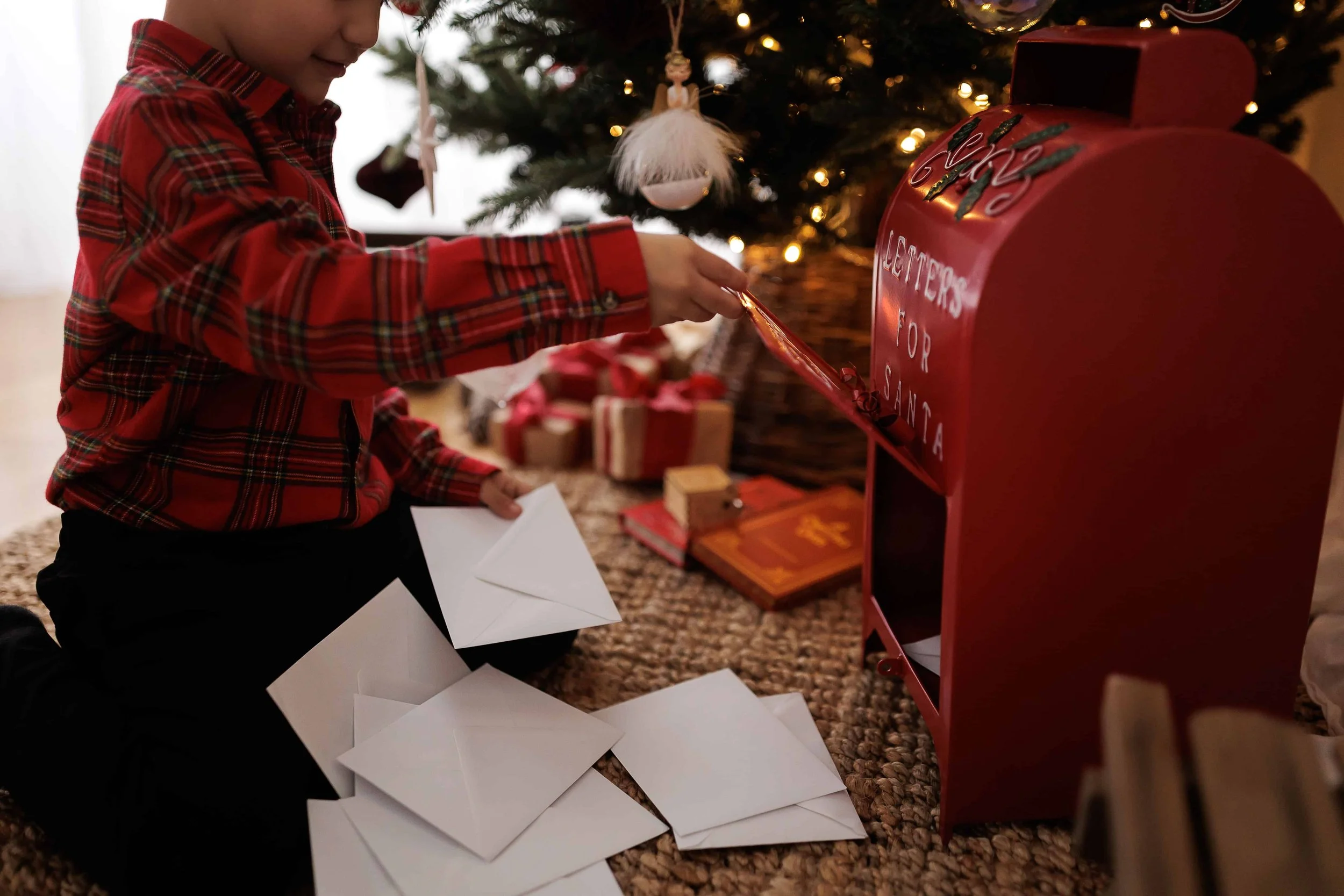 A child in a red plaid shirt sits on a carpeted floor, placing letters into a red Christmas mailbox labeled 'Letters for Santa' under a decorated Christmas tree.