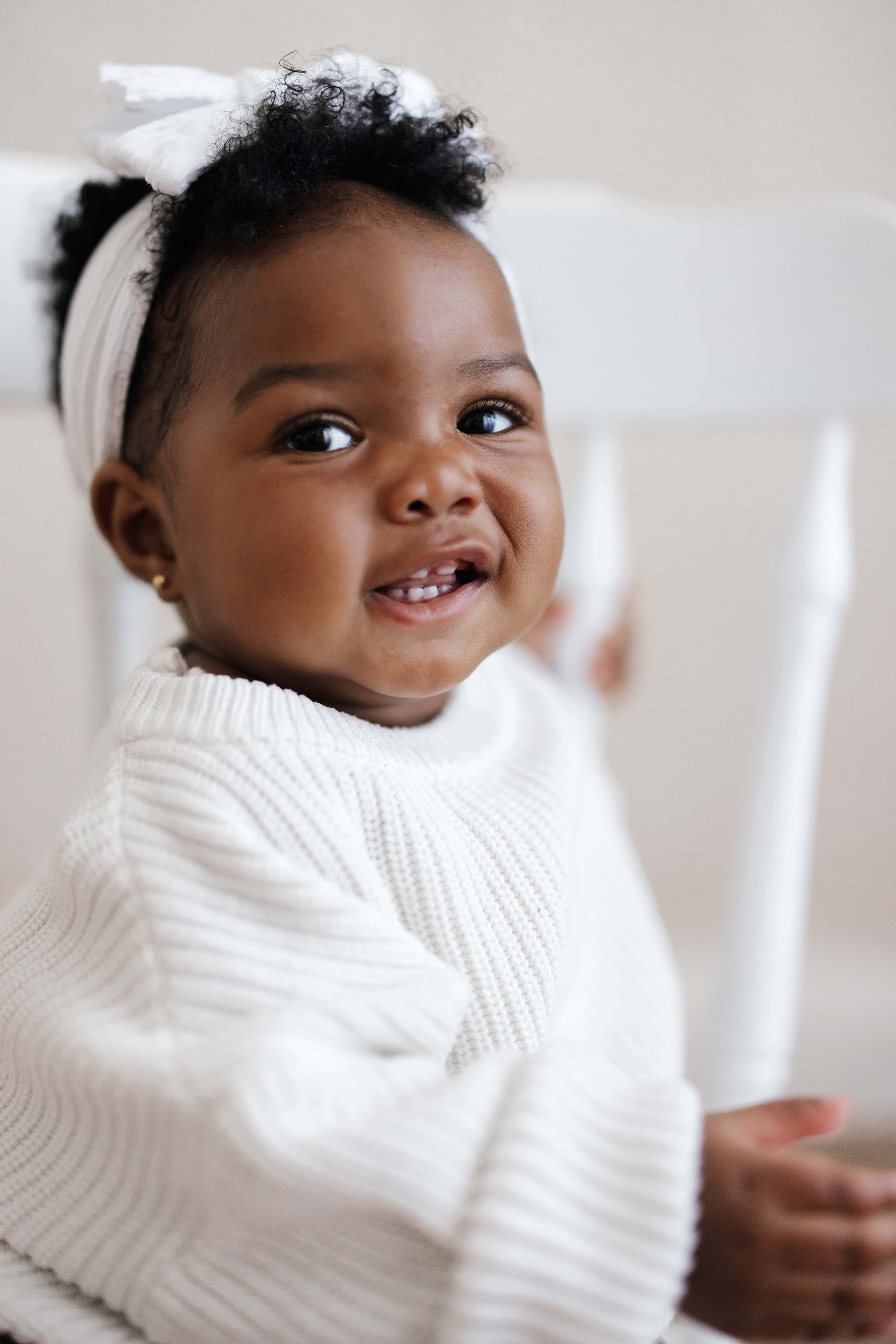 Close-up of a smiling 8 months old baby girl with curly hair tied with a white ribbon, wearing a cream-colored sweater.