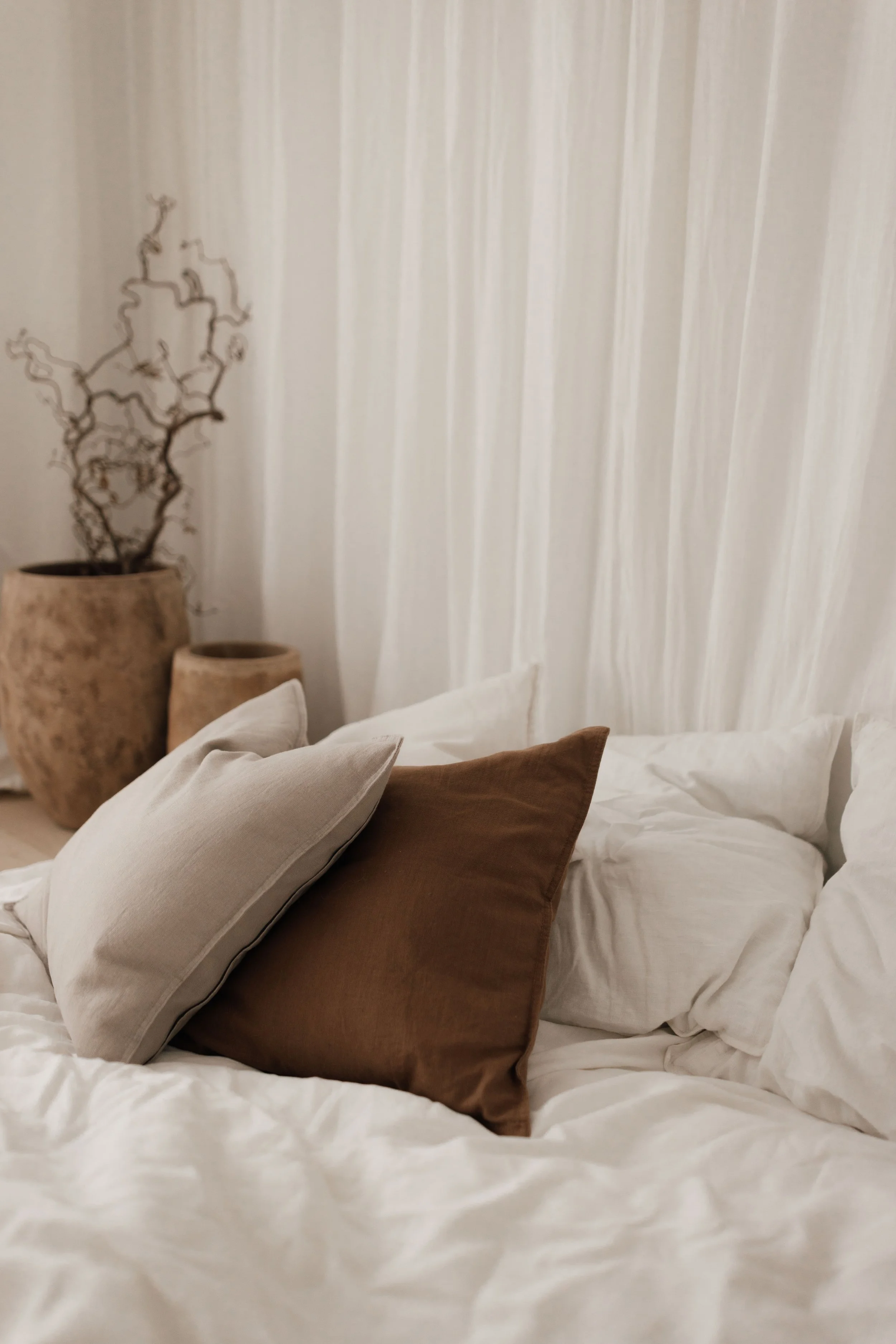 A bed with white bedding and three pillows in beige and brown, with a beige curtain and decorative vases with a plant in the background.