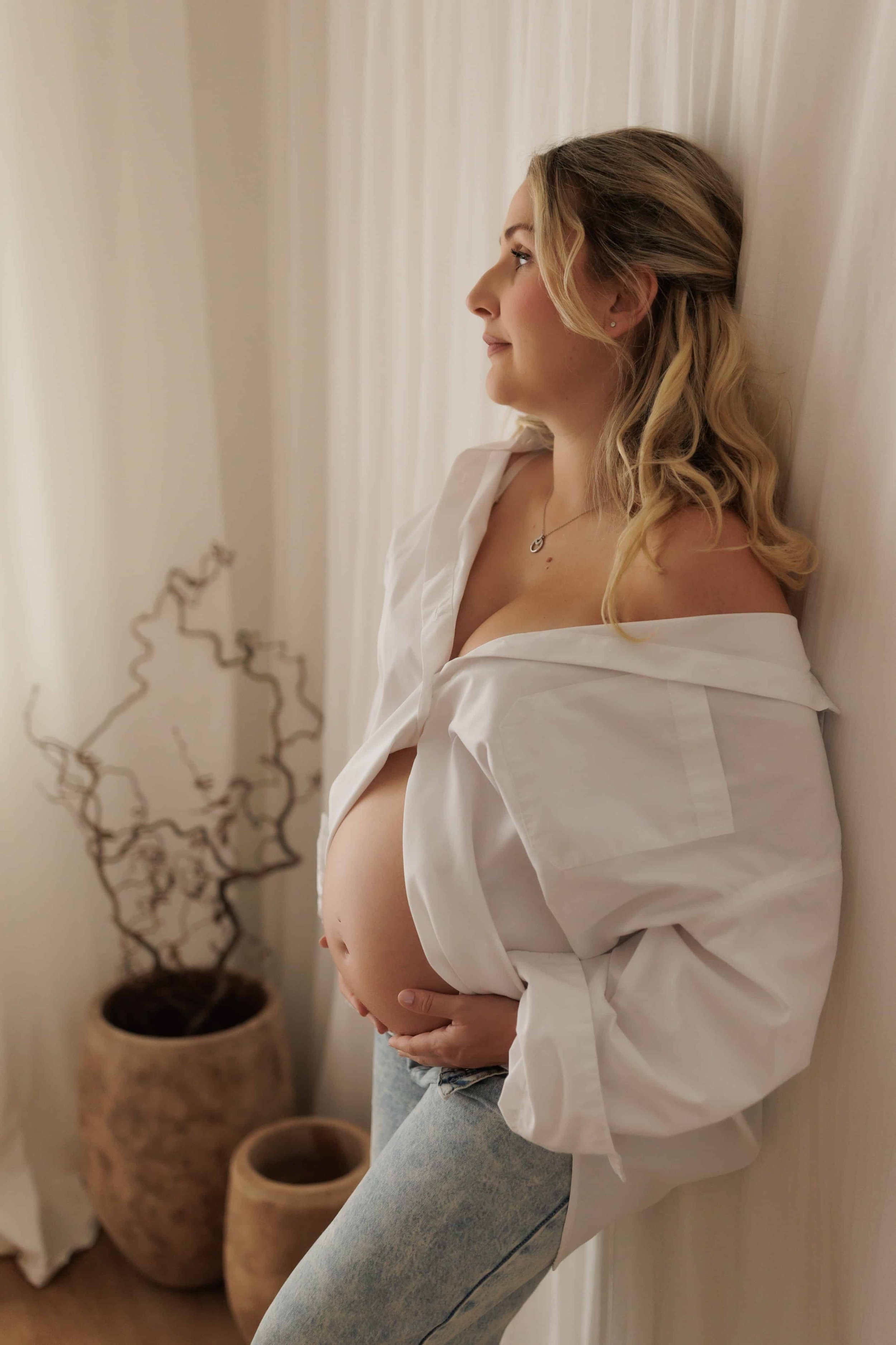 A pregnant woman standing sideways against a wall, wearing a white shirt and light blue jeans, with her hands placed on her belly, and a woman in the background with curly hair. The room has a cream-colored curtain, and a decorative plant with black 