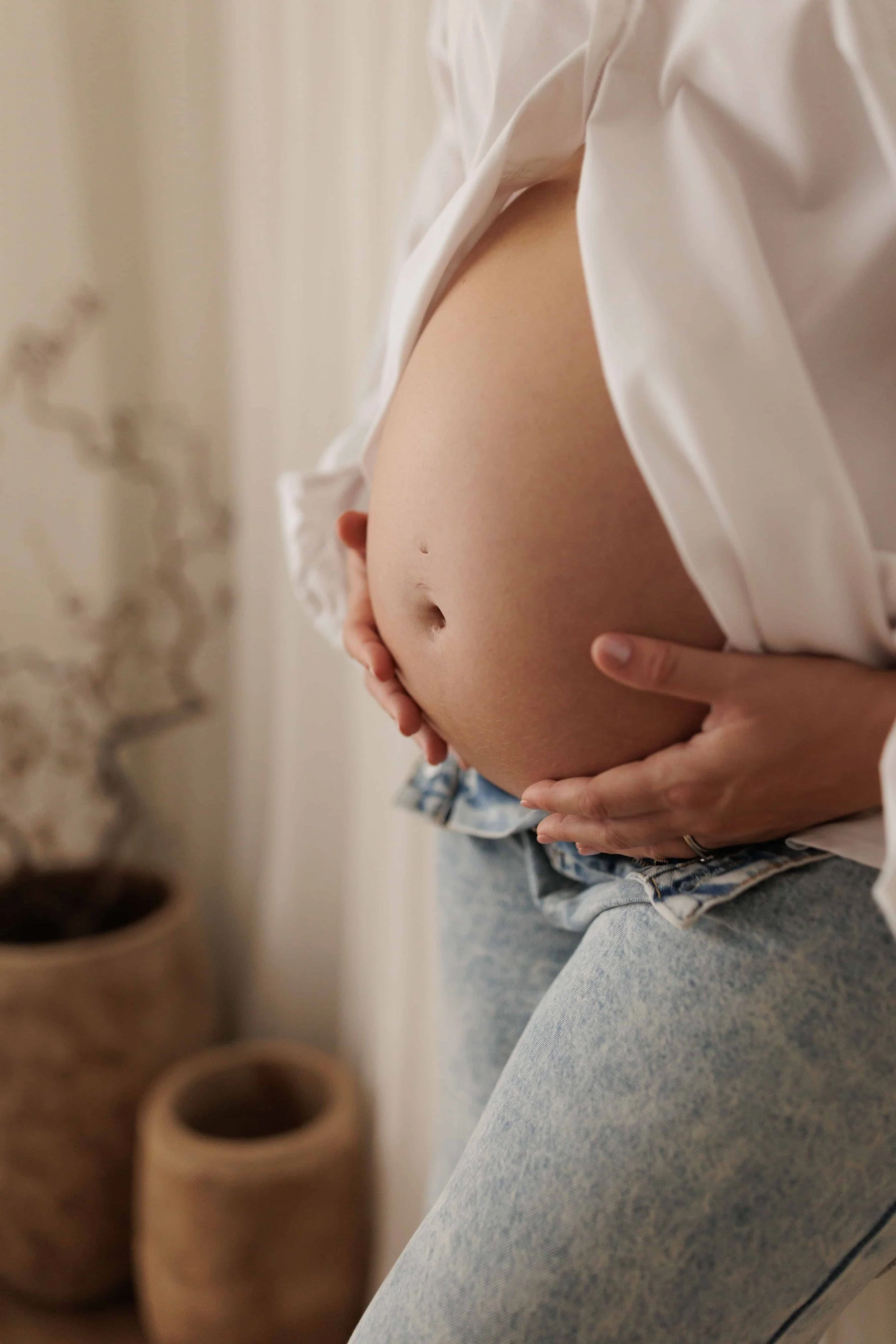 A person wearing a white shirt and light blue jeans is holding their pregnant belly with both hands indoors, with a blurred background showing a white wall and decorative elements.