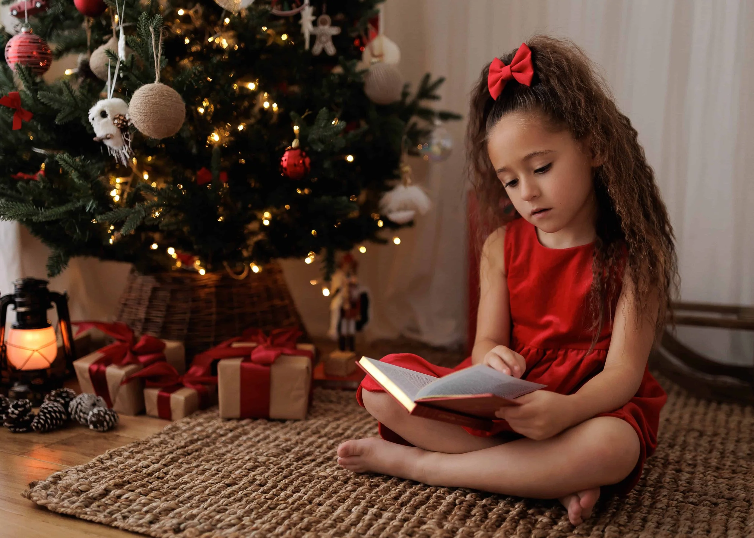 A young girl with long curly hair, wearing a red dress and red bow, sitting on a woven rug, reading a book near a decorated Christmas tree with presents underneath.