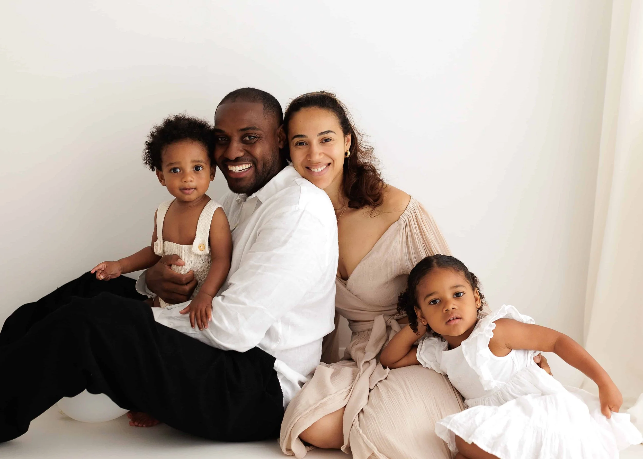 A happy family of four sitting together against a white background, with parents smiling and two young girls dressed in white, one sitting on the woman's lap and the other lying on the floor.