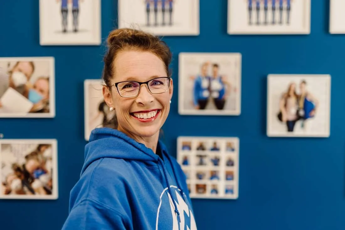A side profile of a woman with short brown hair, wearing a blue sweatshirt, standing in front of a wall of an assortment of pictures.