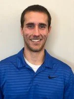 a man with short brown hair and a trimmed beard, smiling at the camera while wearing a royal blue striped Nike polo shirt over a white undershirt, standing against a light beige background.
